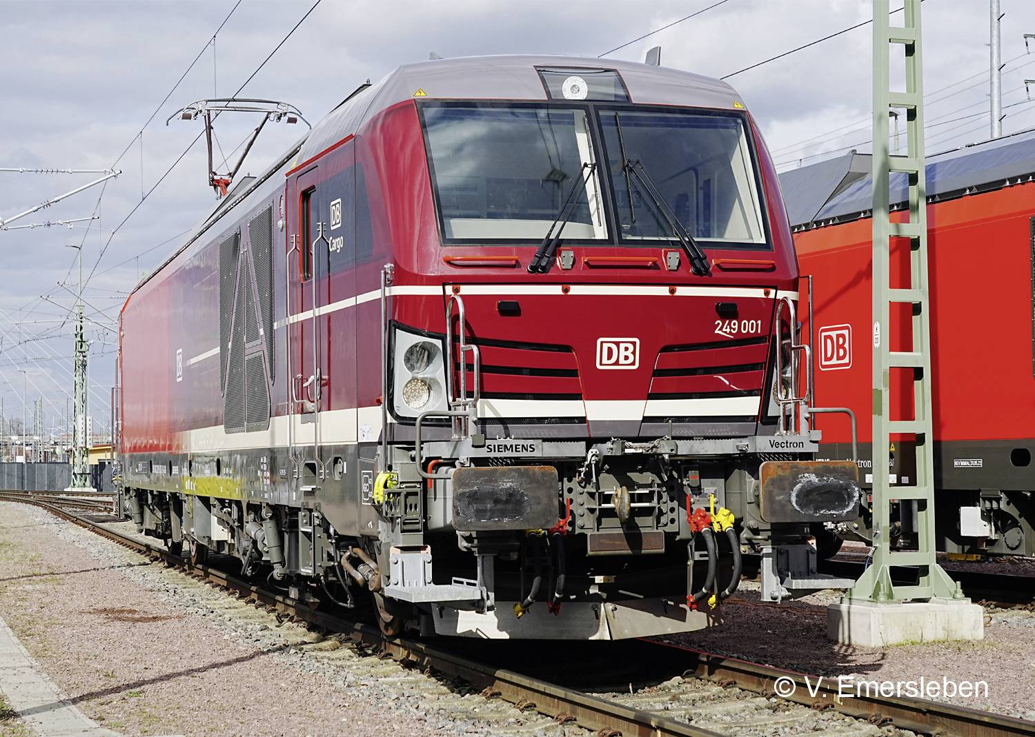 A red locomotive of Deutsche Bahn is standing on a railway track. Overhead power lines are located to the left.