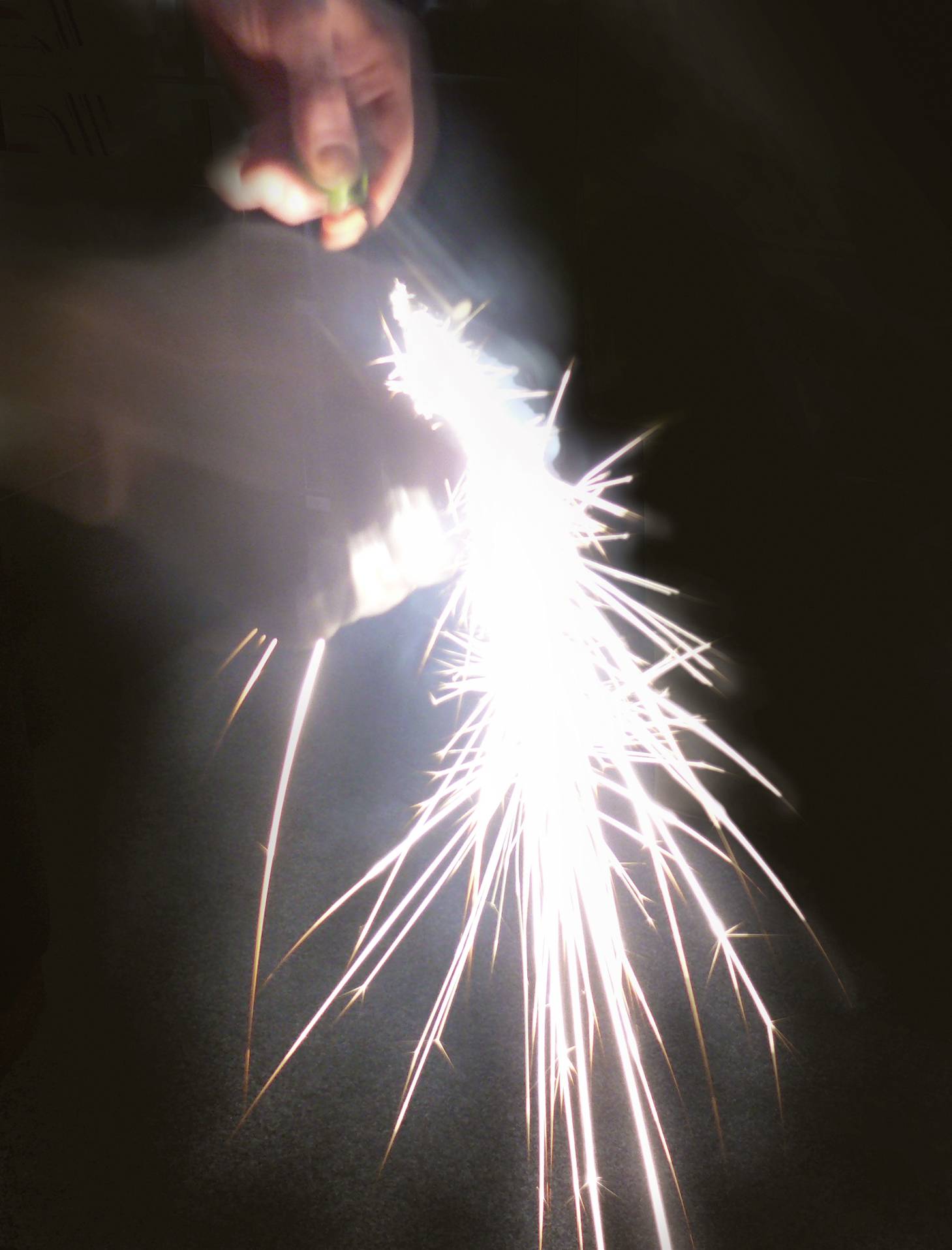 A hand holds a sparkler, casting bright sparks. The background is dark, causing the sparkler's light to stand out vividly.
