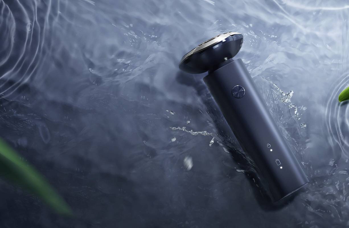 An electric razor floats in the water. Dark casing with a silver head. Surrounding environment with water surface and plant elements.