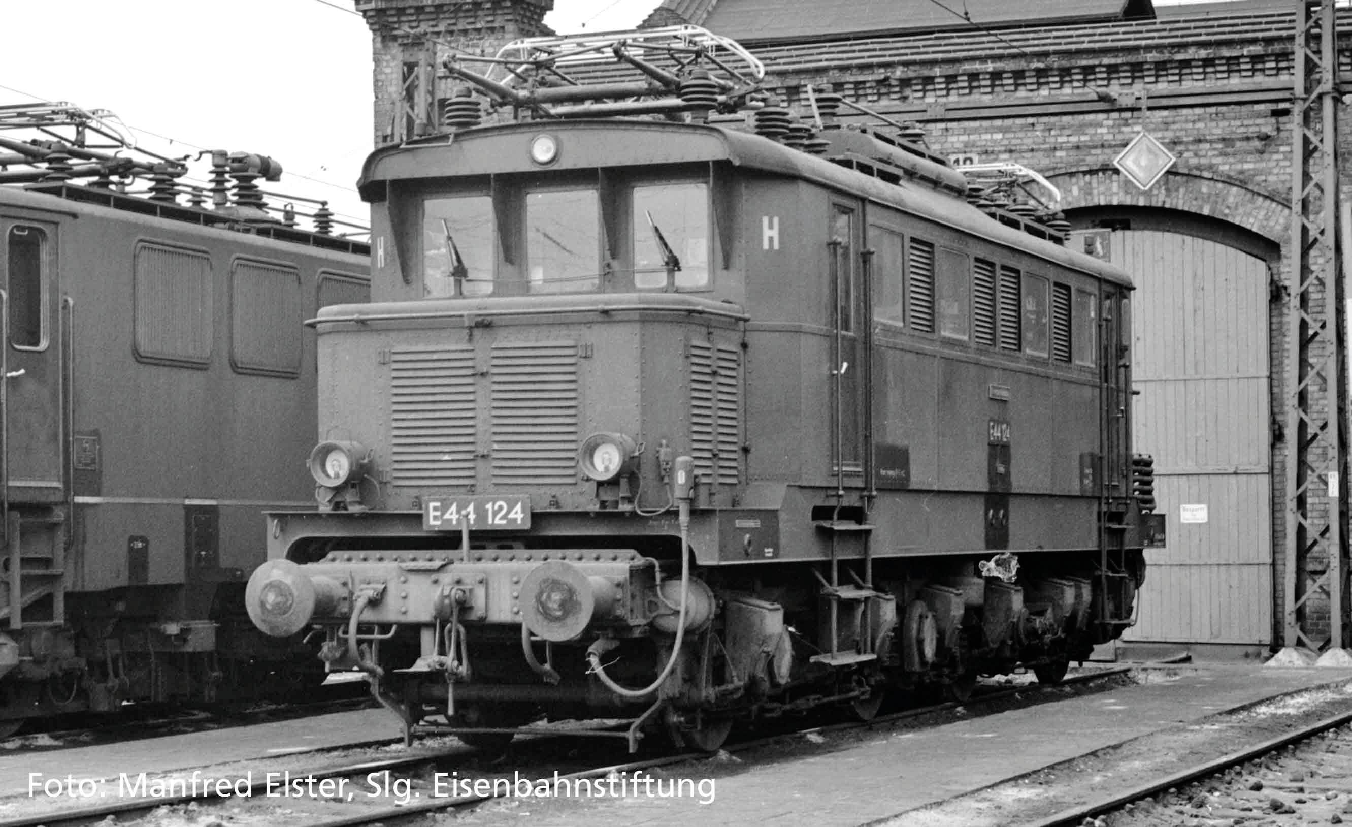 A historic electric locomotive stands in front of an old railway depot. It bears the designation 'E41 124'.