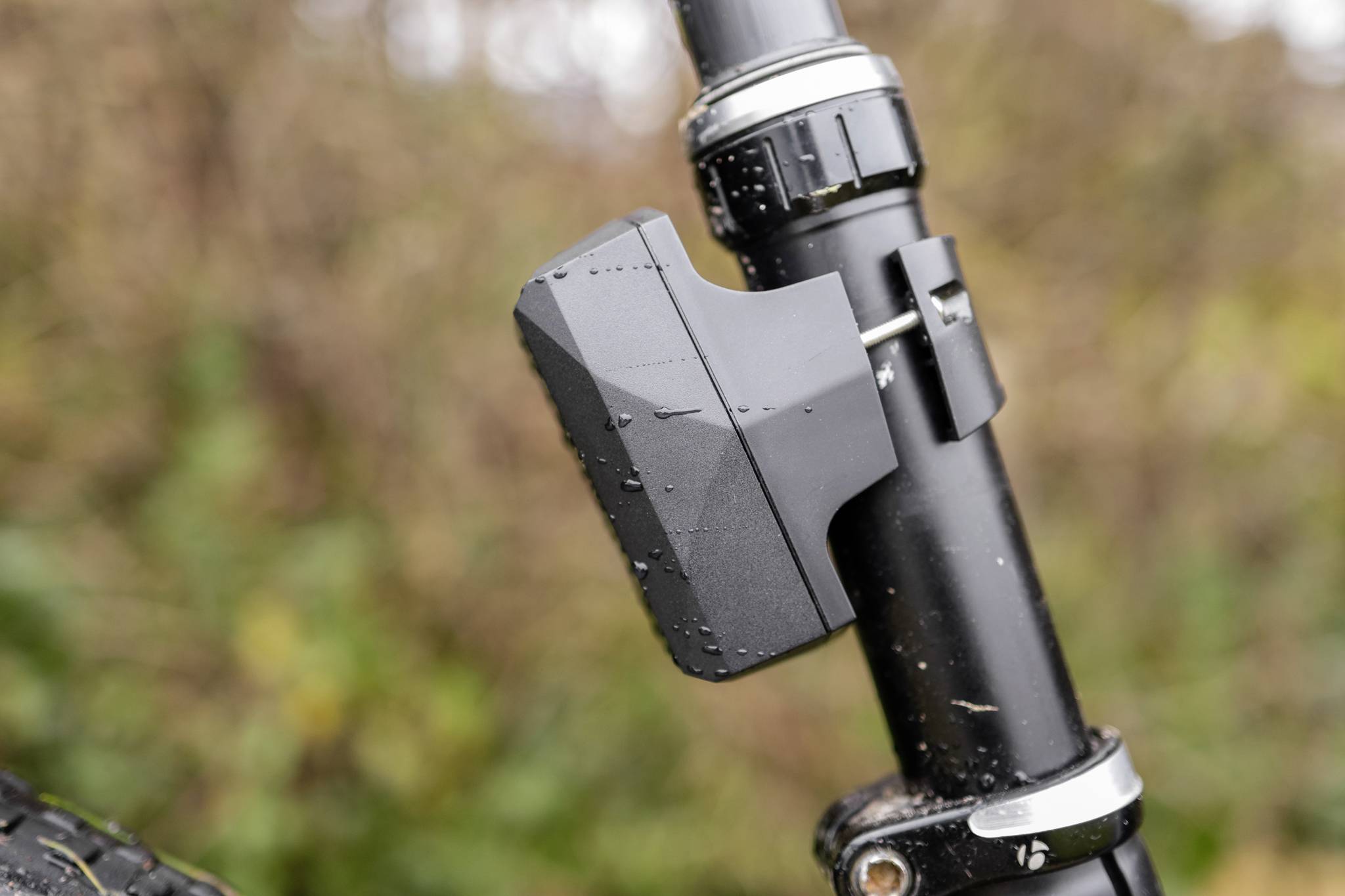 A black bicycle light is attached to the seat post of a bicycle. Blurred green plants are visible in the background.
