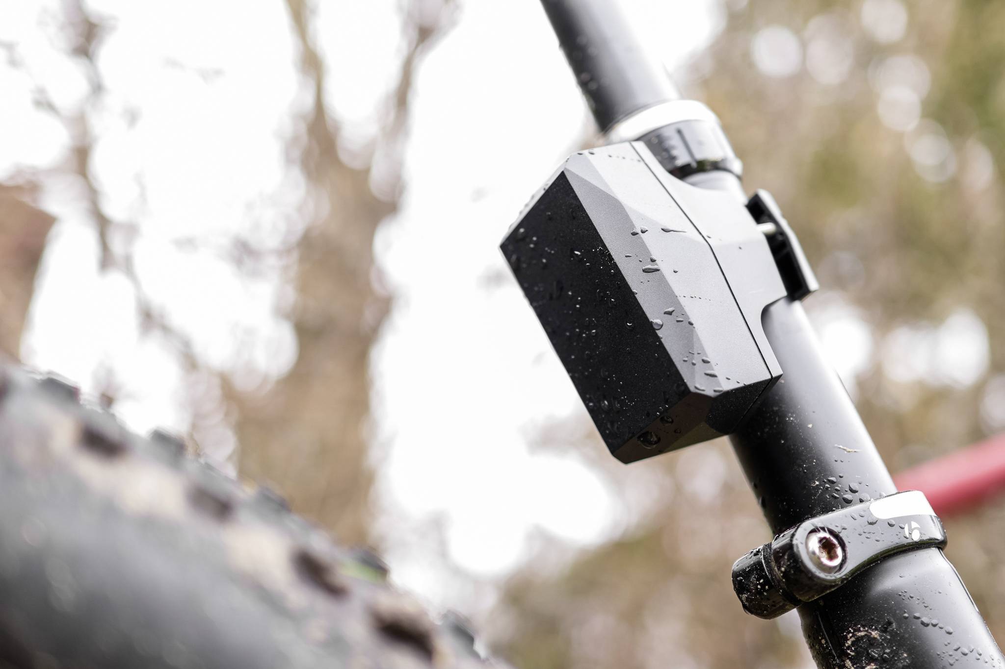 Close-up of a bicycle frame with a mounted box and mud-splattered tyre; autumn leaves blurred in the background.