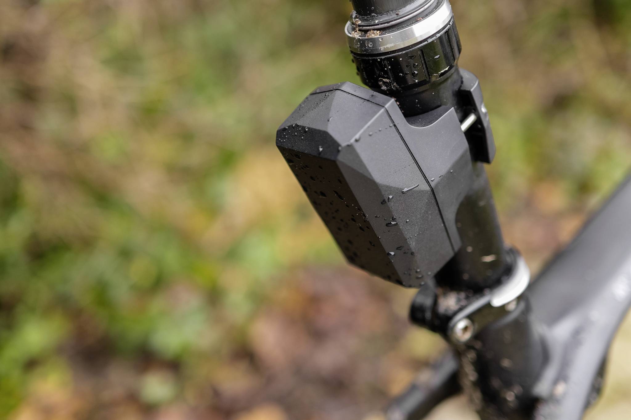 Close-up of a bicycle component with an electronic device attached to the frame. The device is speckled with raindrops. Background blurred, with autumnal colours.