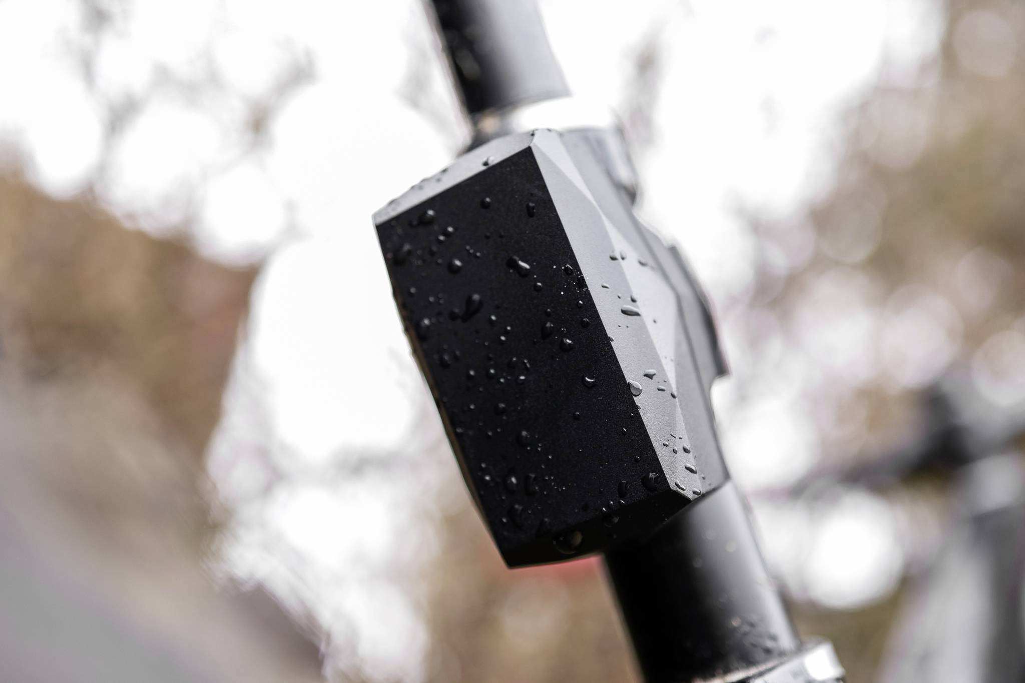 A black device covered in water droplets is attached to a pipe or handlebar. Blurred tree branches can be seen in the background.