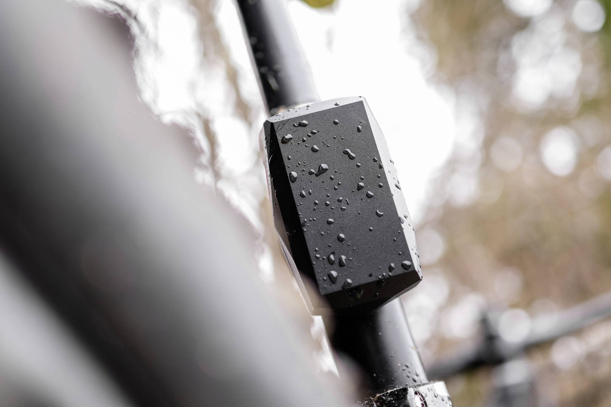 A black, rain-soaked object is attached to a pole. Blurred background with trees. Weather-resistant device outdoors.