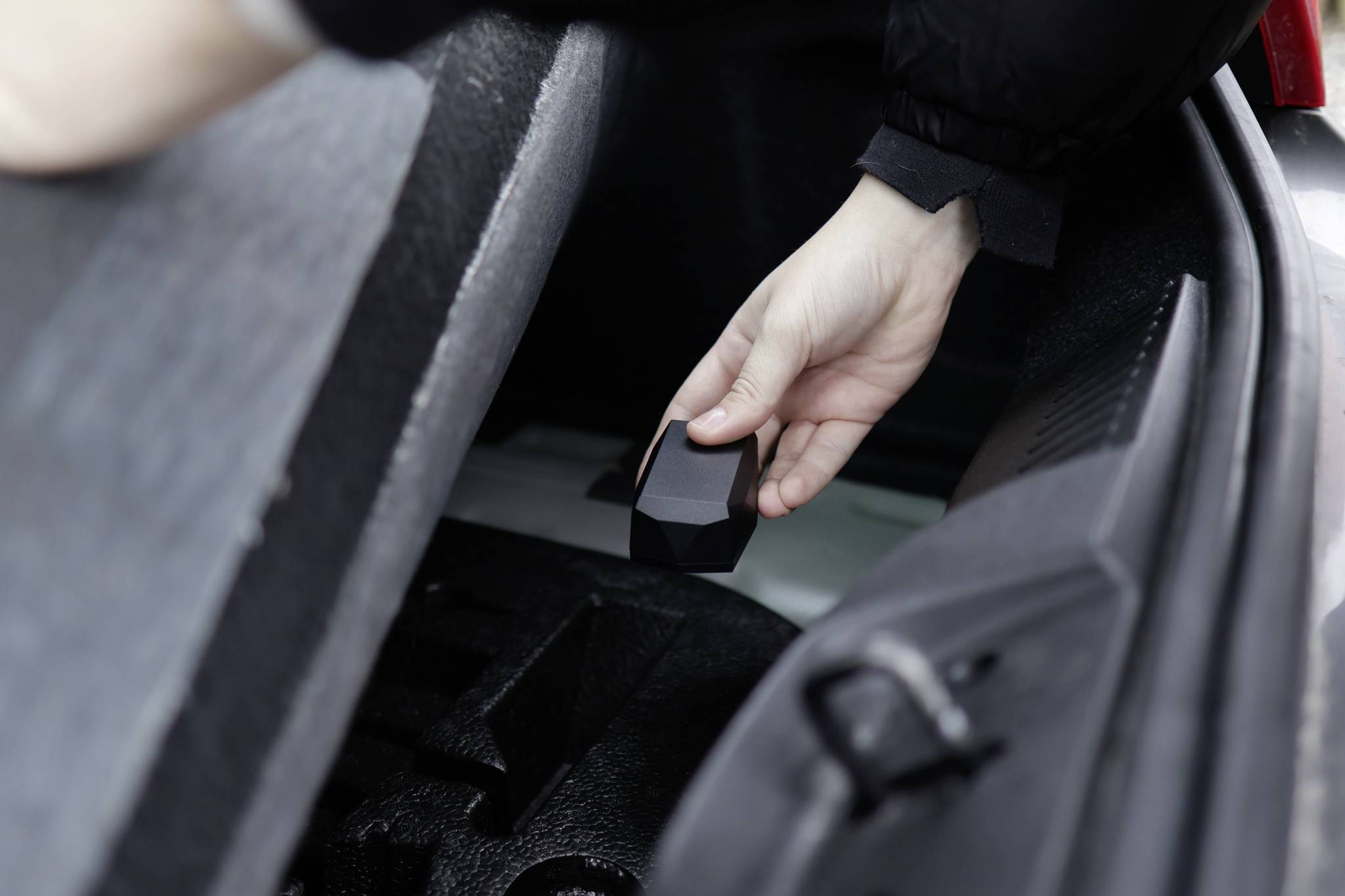 A hand reaches for a black object in the boot of a car. The scene depicts the storage or removal of an item from the luggage compartment.