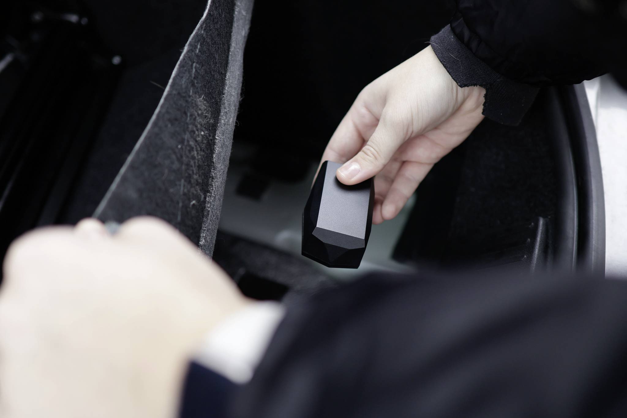 A person is holding a small, black, geometric object in their hand over an open car boot.