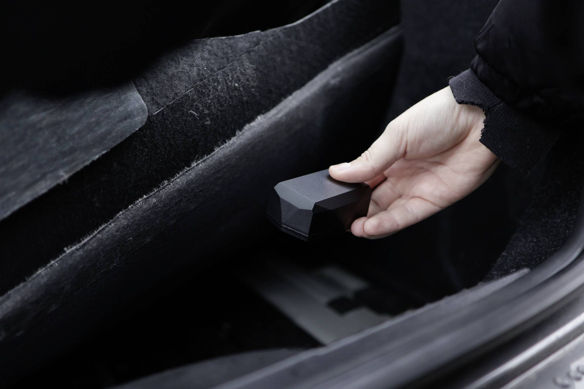 A hand is holding a small black object in the boot of a car. The object appears to be an accessory or tool.