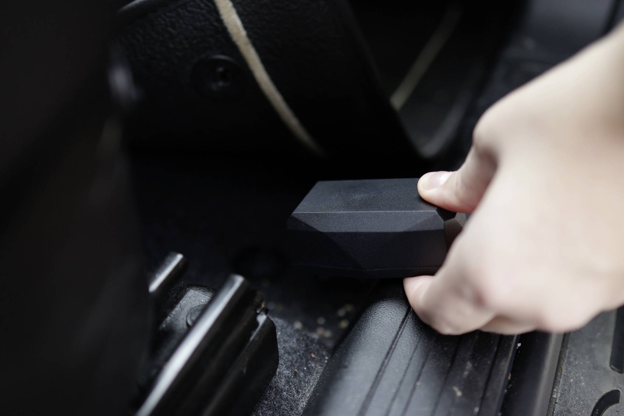 A person is holding a small, black device in their hand and placing it in the footwell of a vehicle, close to the seat frame.