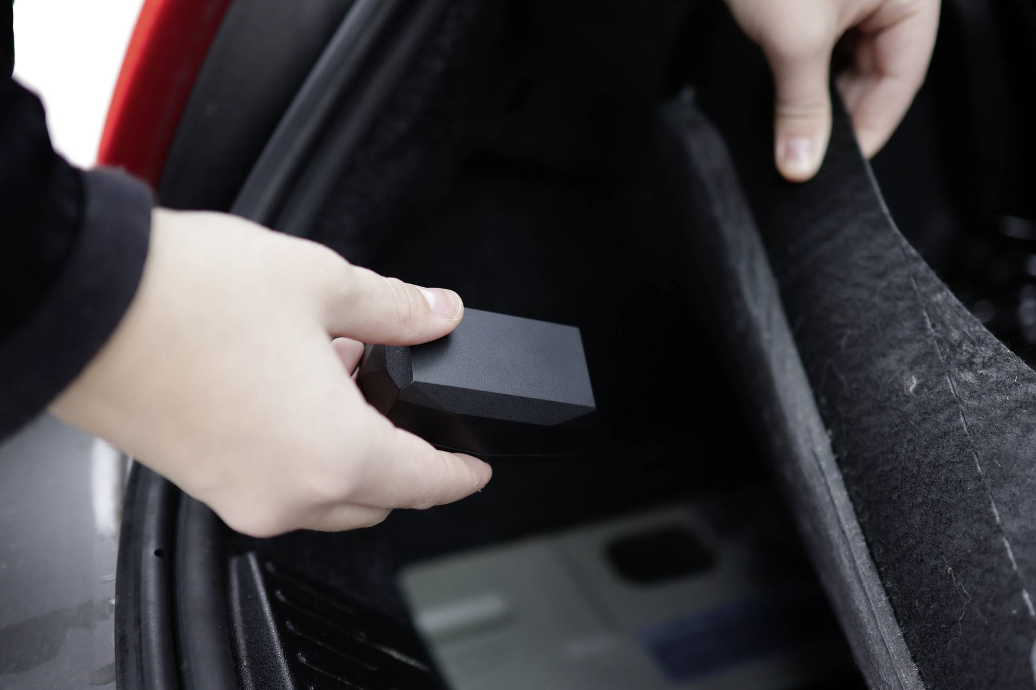 A person places a small, black device in the boot of a car. The device could be a GPS tracker or a similar electronic device.