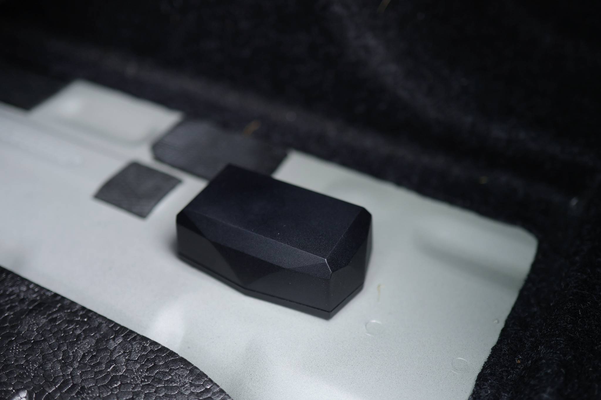 A black, rectangular object with bevelled corners lies on a grey surface, surrounded by black and grey areas.