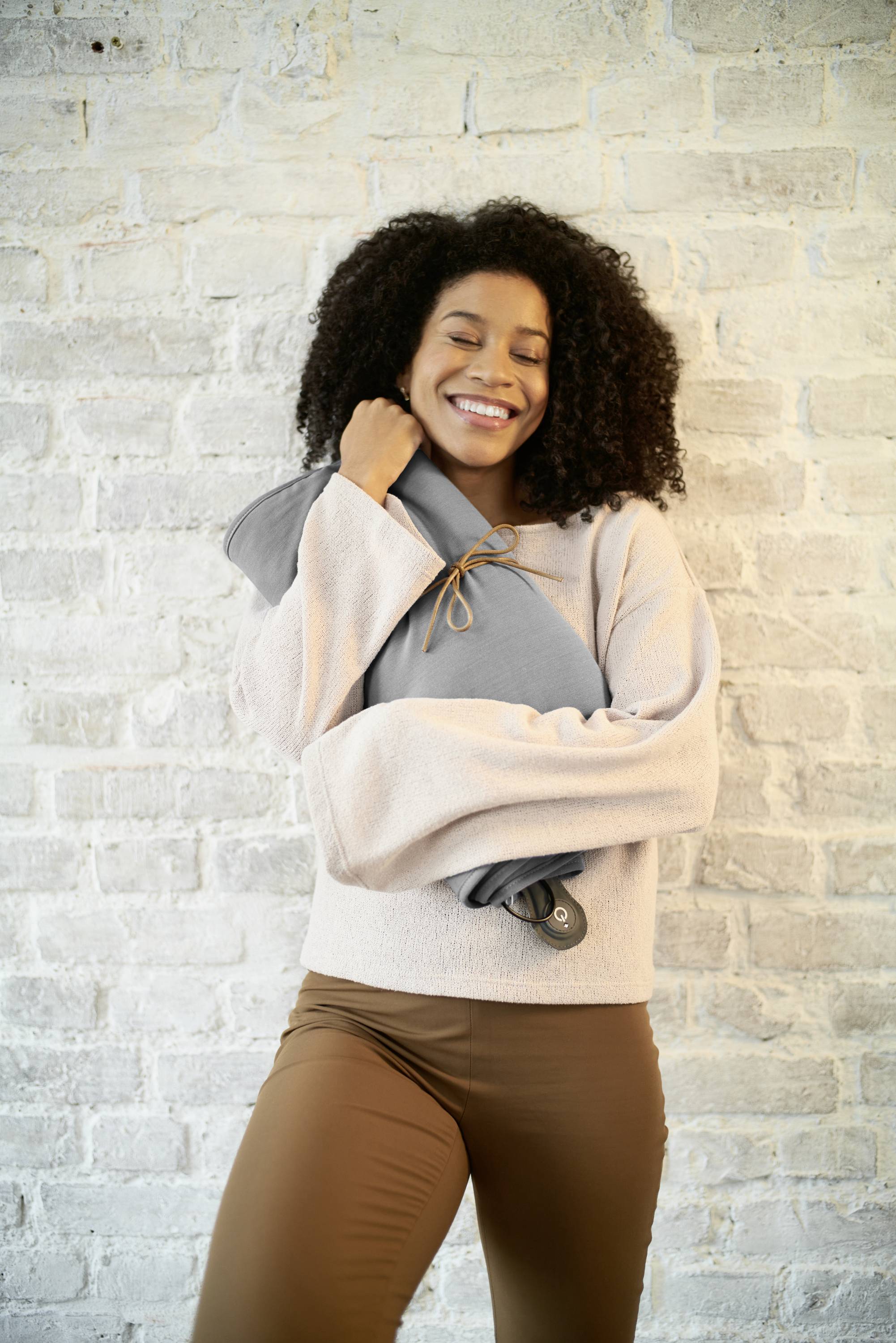 A woman in beige clothing smiles and holds a warm, grey cushion in front of a white brick wall.