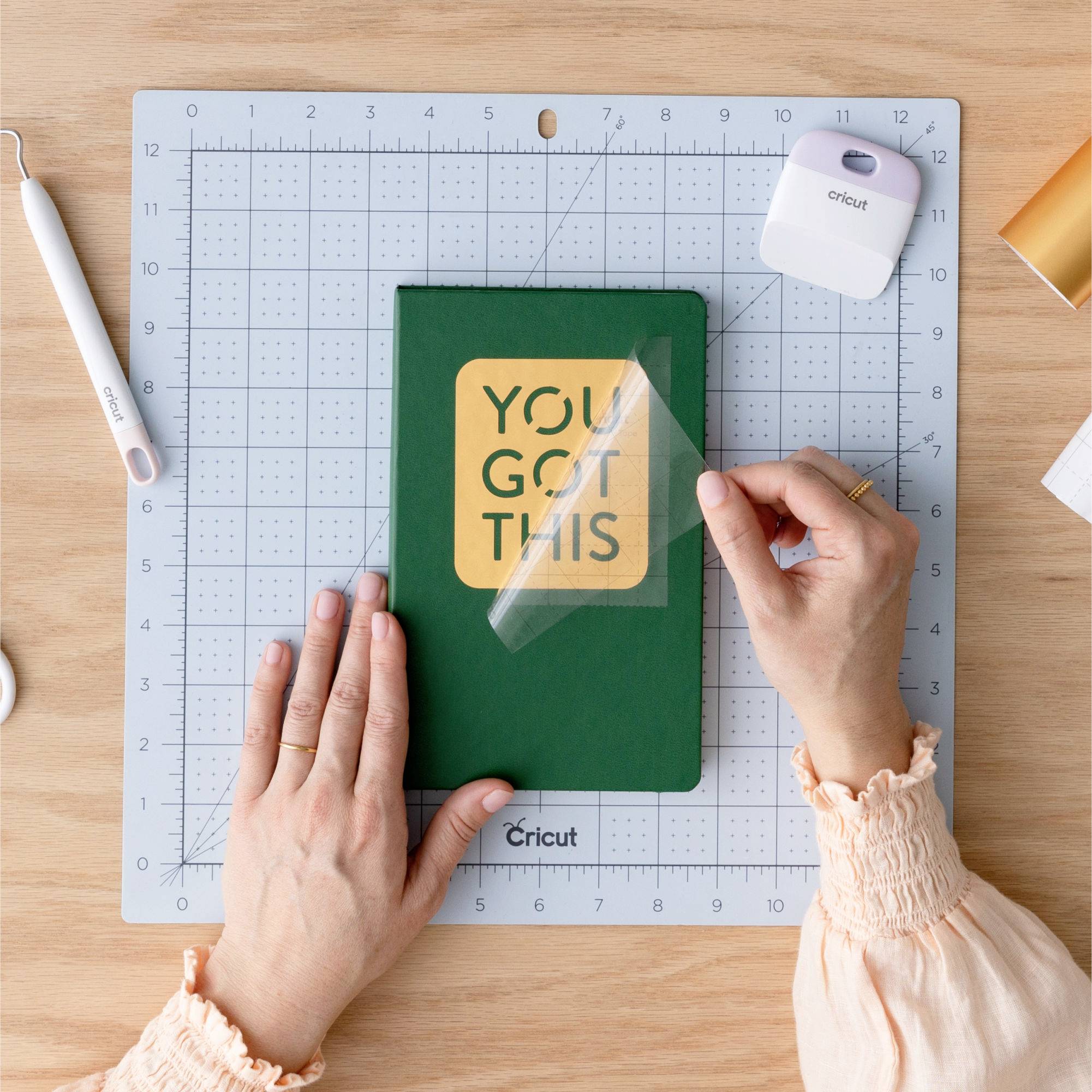 A person is sticking a sticker saying 'YOU GOT THIS' onto a green notebook. Craft tools and materials are scattered around.