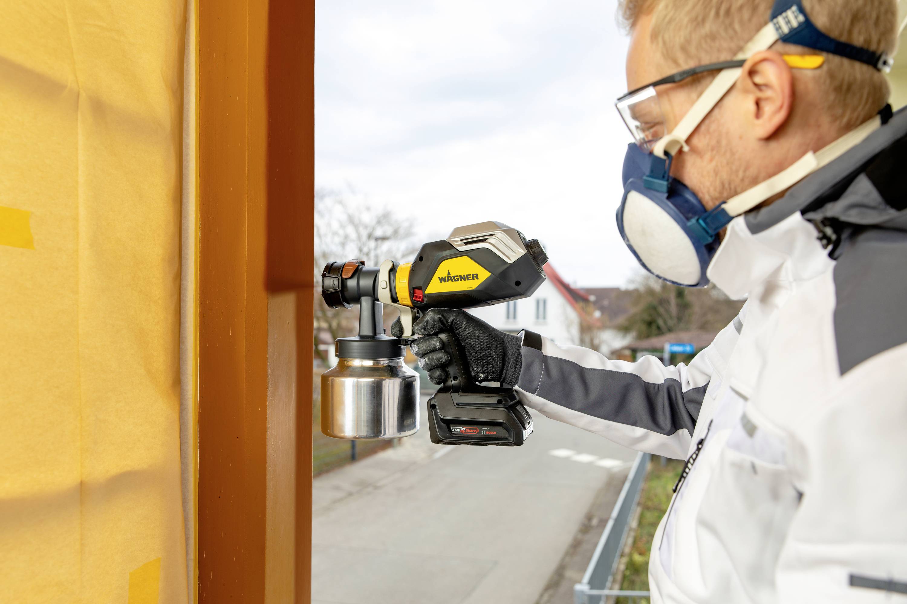 A man in safety equipment is spraying paint with a spray gun onto a wooden frame outdoors.