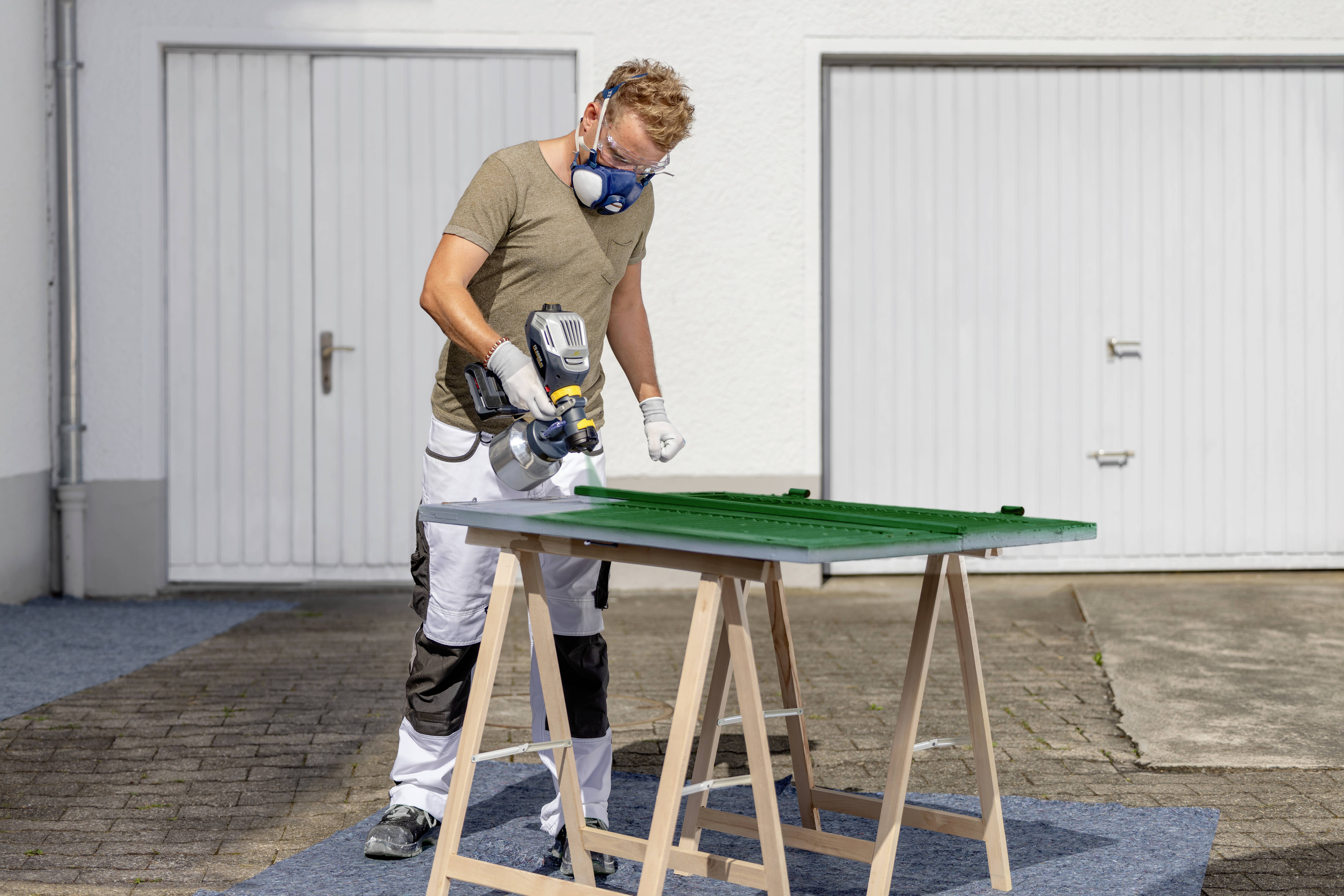 A man in protective clothing is painting a green wooden board on an easel in front of a white garage door.
