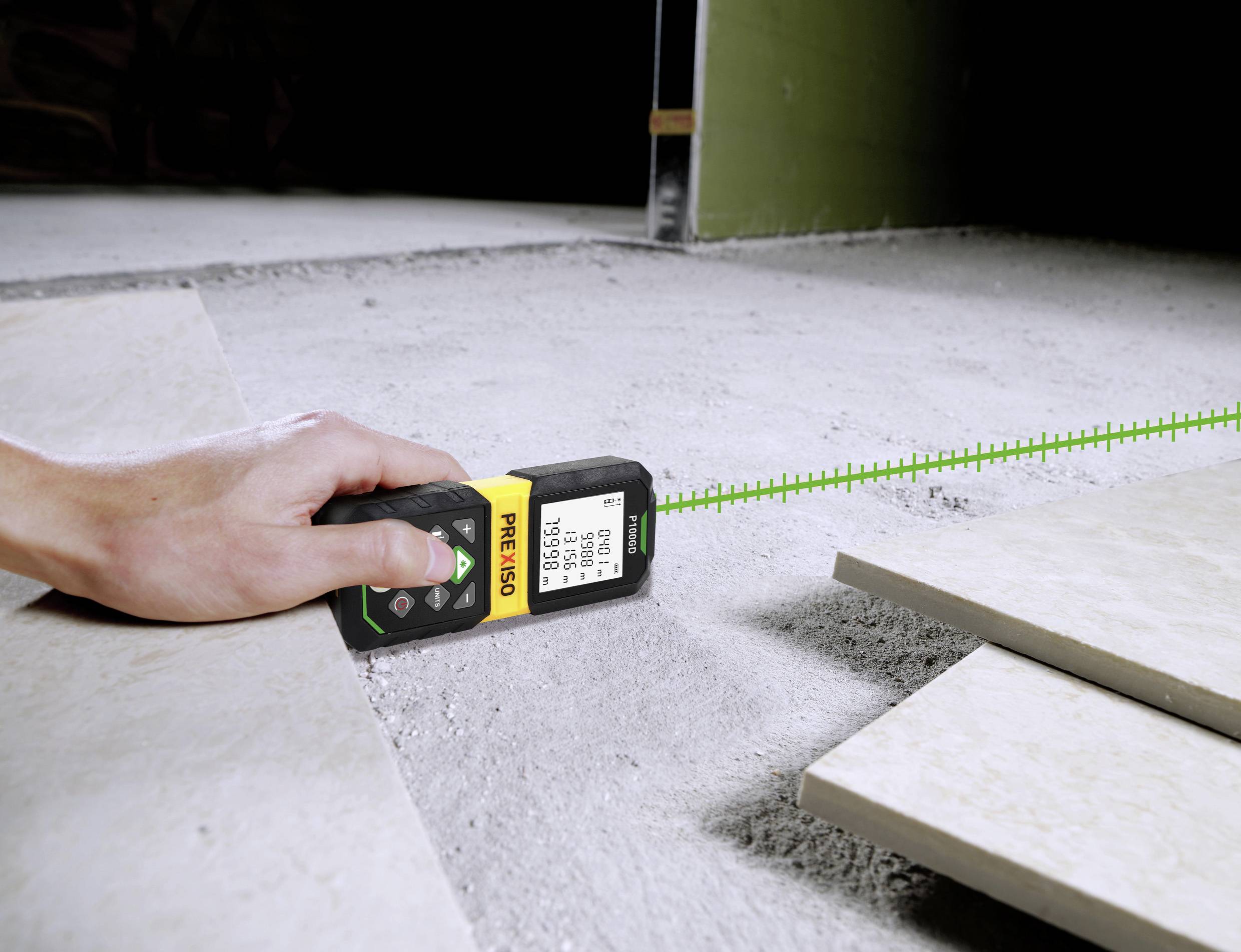 A hand is holding a laser measuring device on a concrete floor, measuring the distance to floor tiles. The device displays a measured distance.