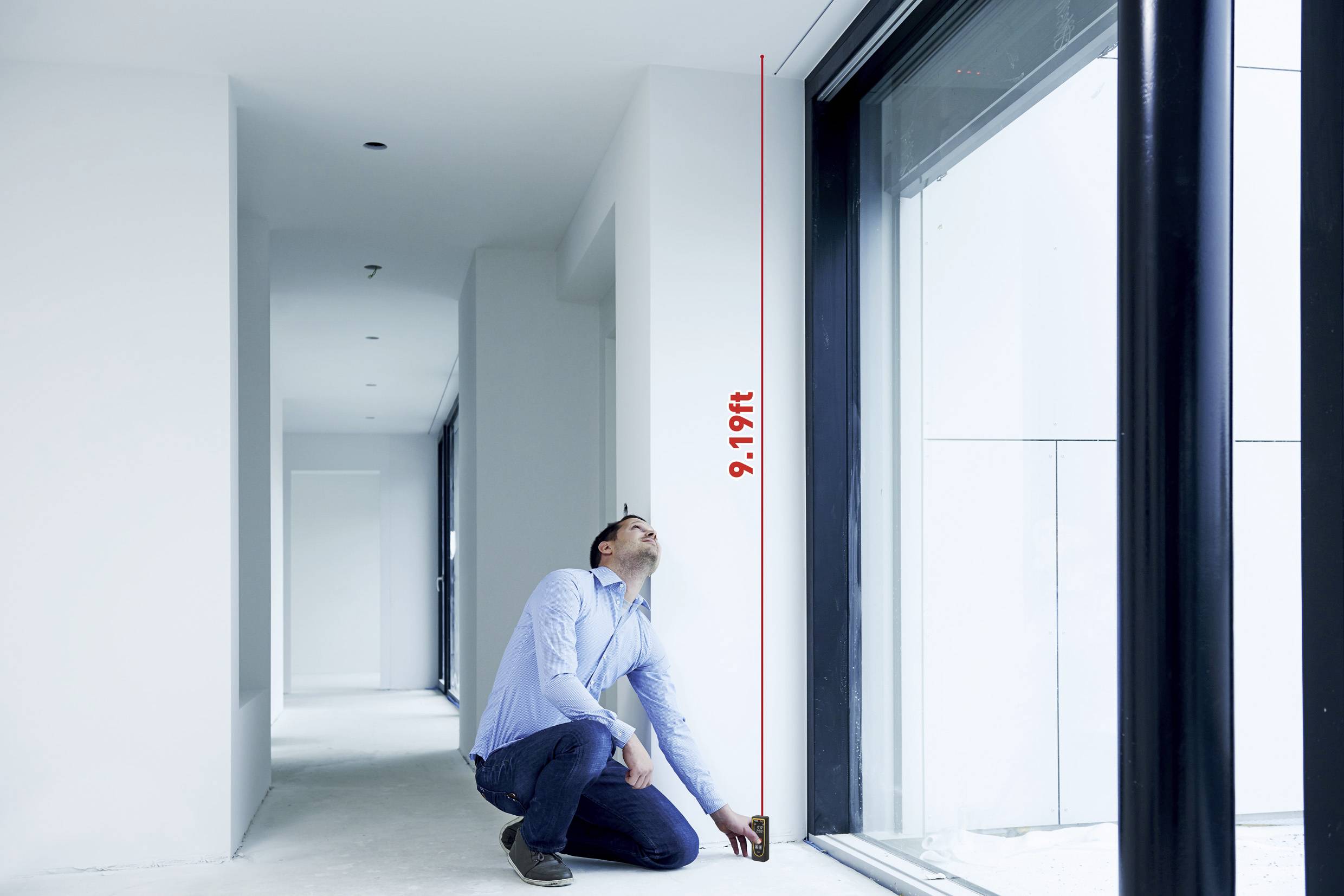 A man is kneeling in a bright room and measuring the height of a large window with a tape measure. The height is 9.19 feet.