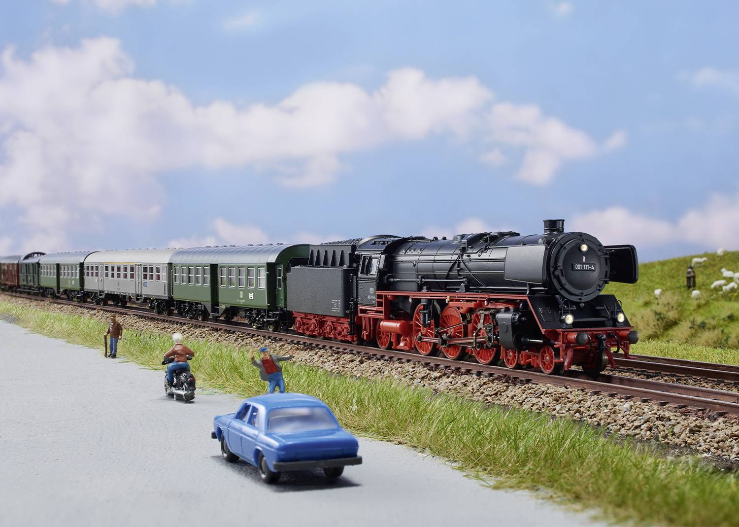 A black steam train travels along tracks through a green landscape under a blue sky. In the foreground of the image, a car and people can be seen.