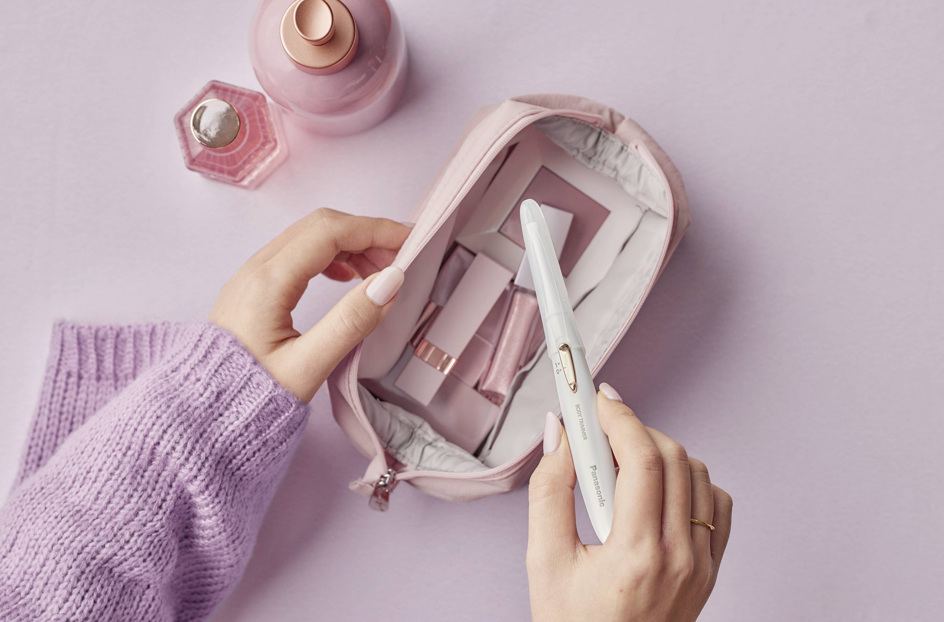 A woman packs cosmetic items into a pink toiletry bag, against a lilac background. Two perfume bottles are positioned in the top left corner.