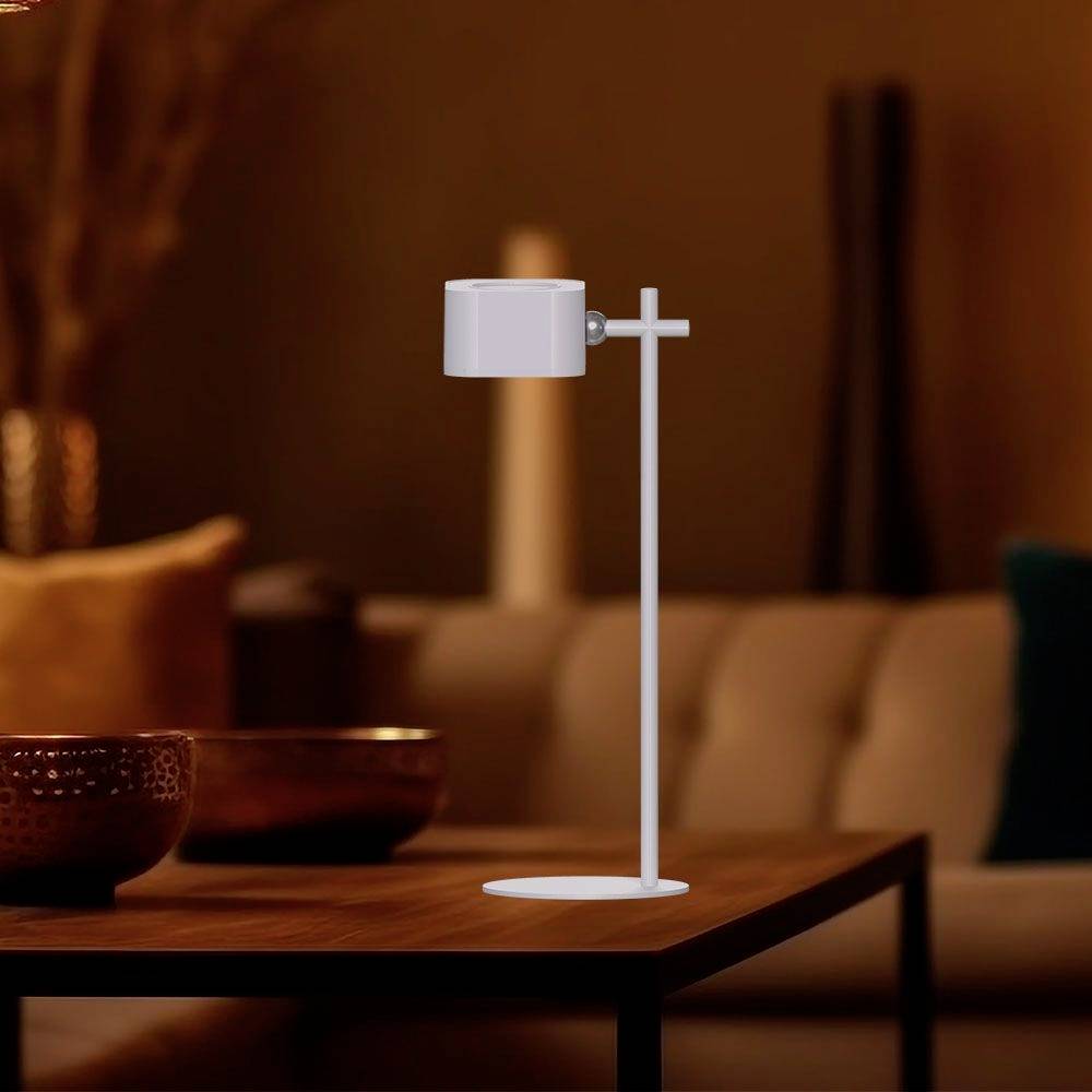 A modern, white LED table lamp sits on a wooden table in a cosily lit living room with a sofa in the background.