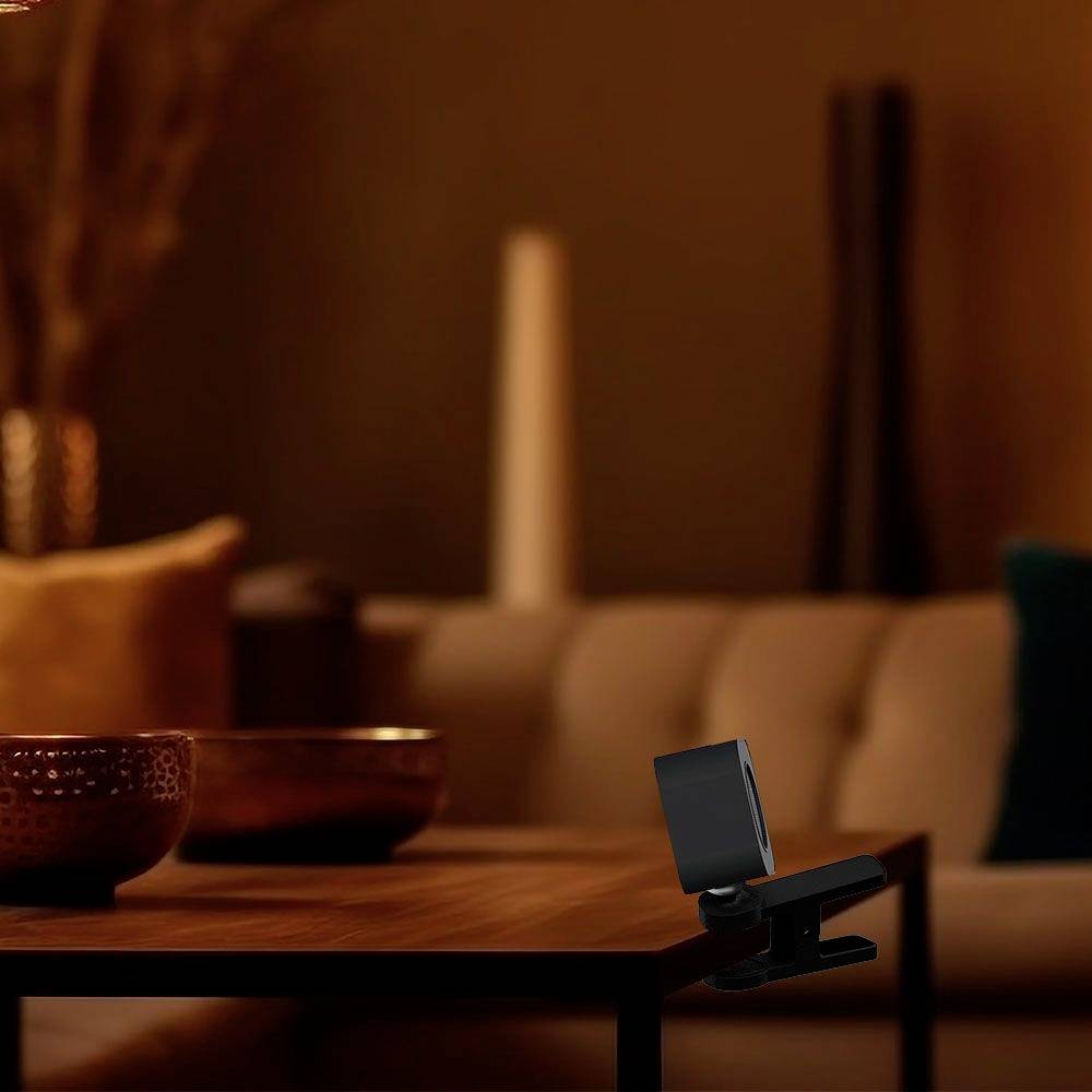 A modern webcam sits on a wooden table in front of a blurred living room background with beige and navy cushions.