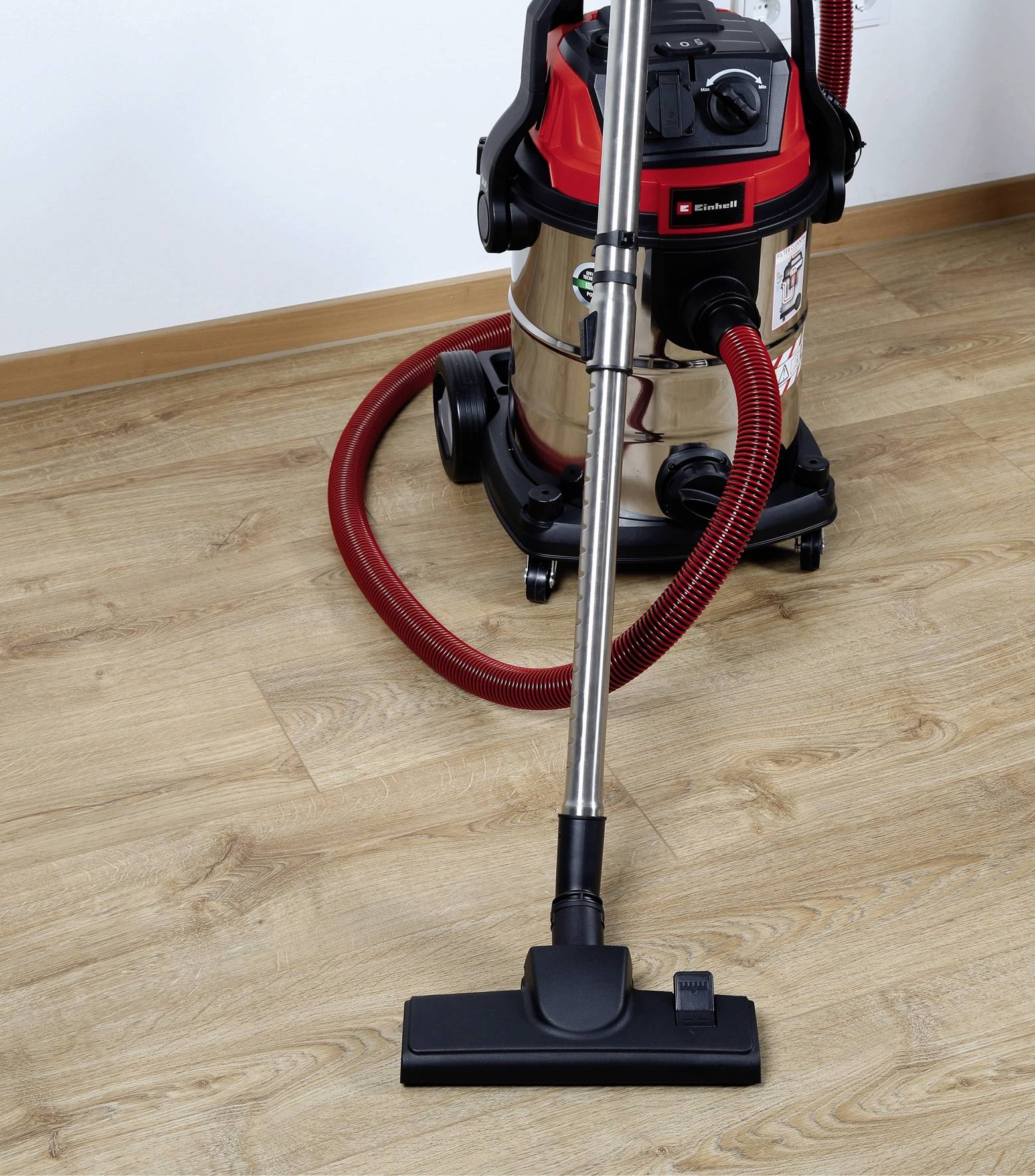 A powerful vacuum cleaner with a red hose stands on a wooden floor, ready for cleaning.