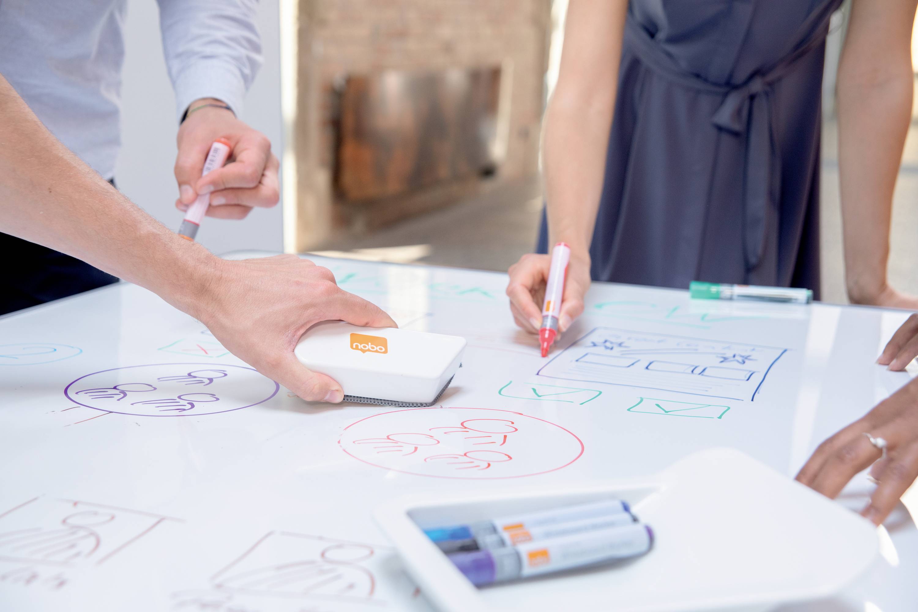 People are drawing with coloured pens on a white table being used as a whiteboard. An eraser is in the foreground.