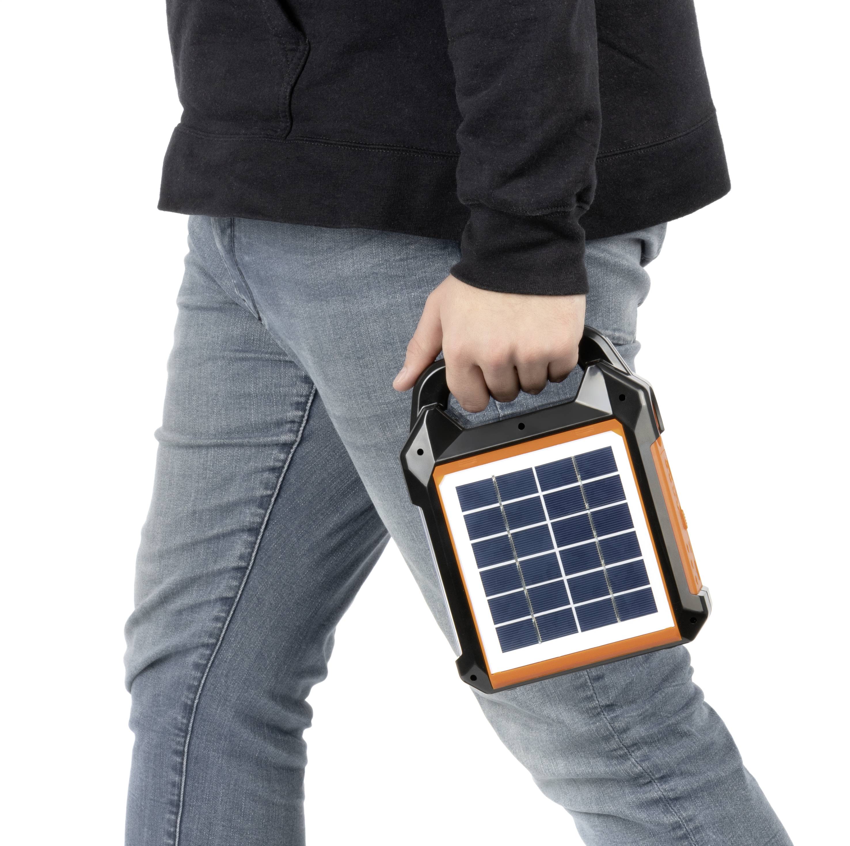 A person in jeans is carrying a portable solar generator in their hand. The device has a solar panel on its front.