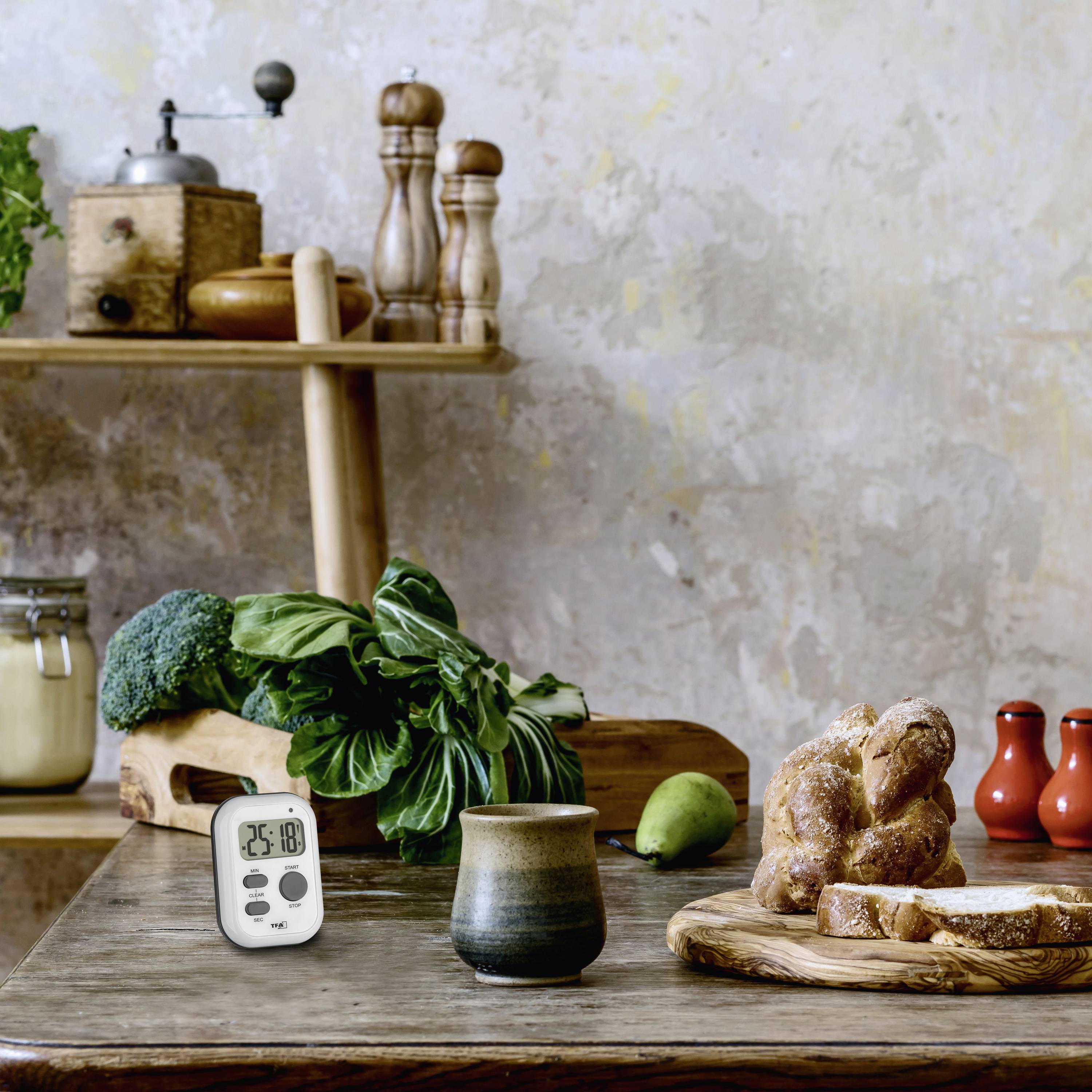 A table with fresh bread, broccoli, a timer and a mug, with a rustic kitchen environment and spice mills in the background.