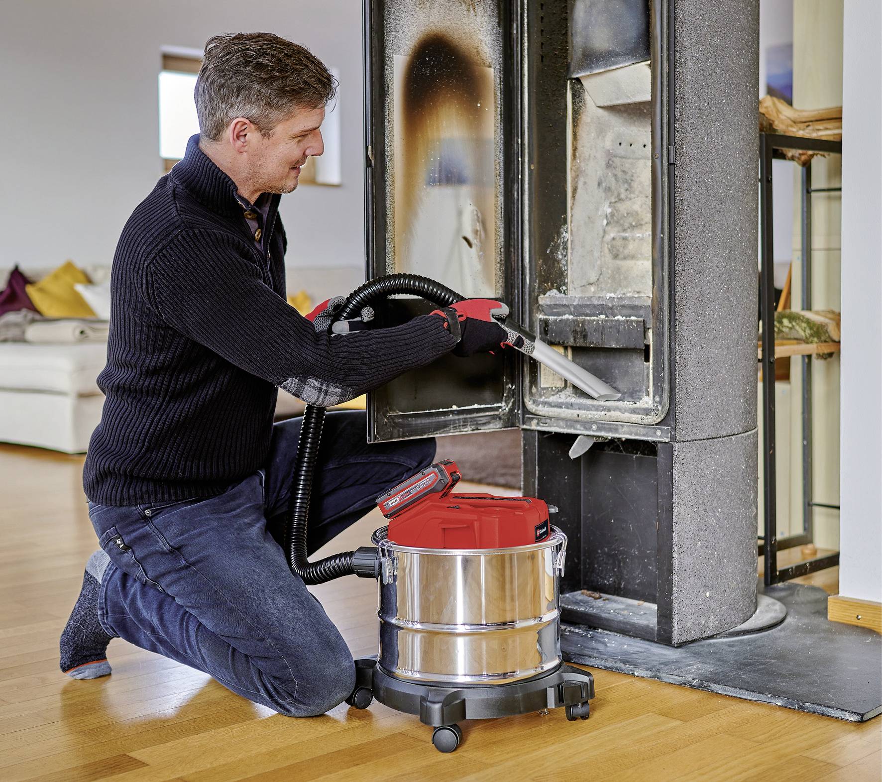 A man is cleaning a chimney with an ash vacuum. He is wearing black clothing and gloves, with the living room visible in the background.