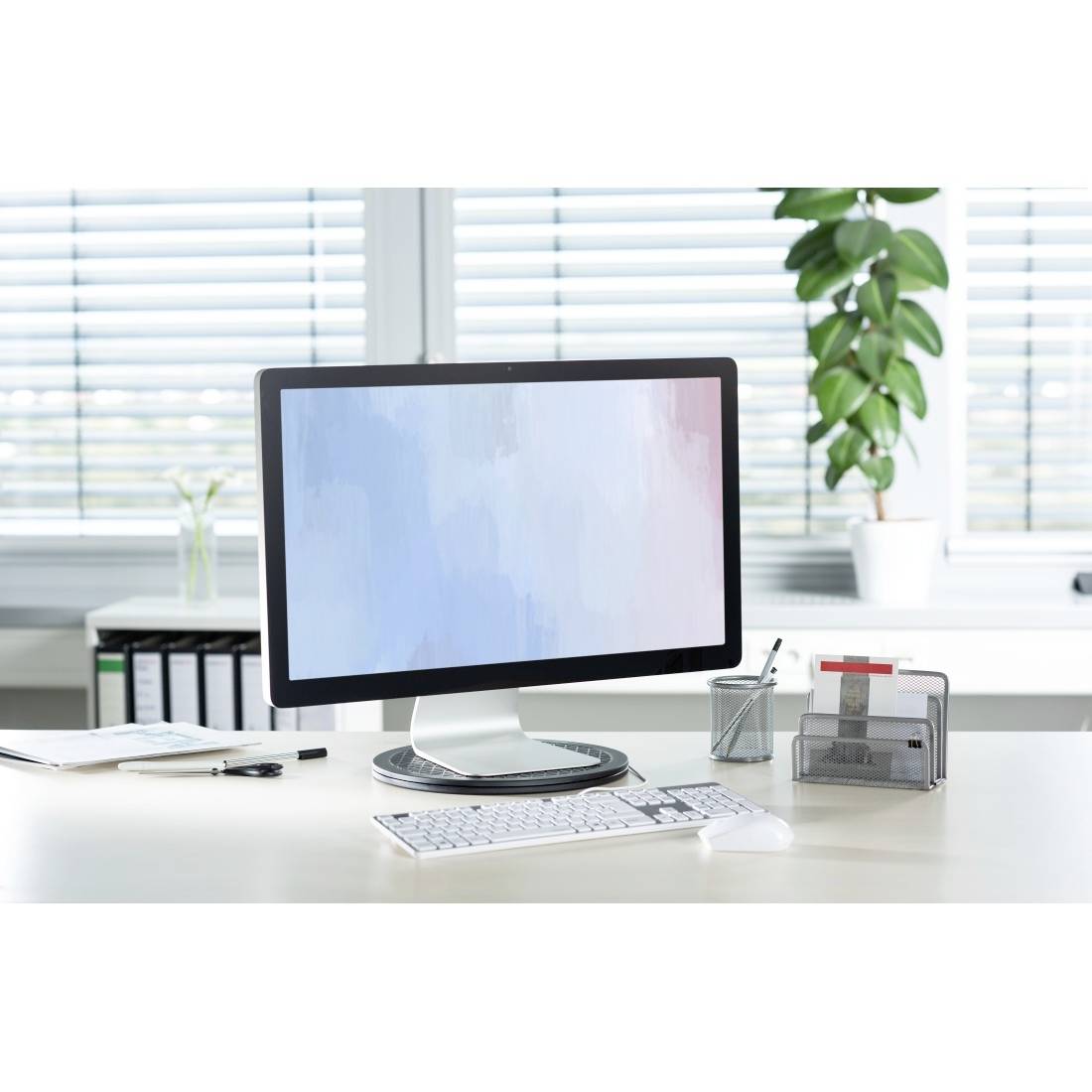 A modern computer monitor on a white desk, surrounded by office supplies and a potted plant in the background.