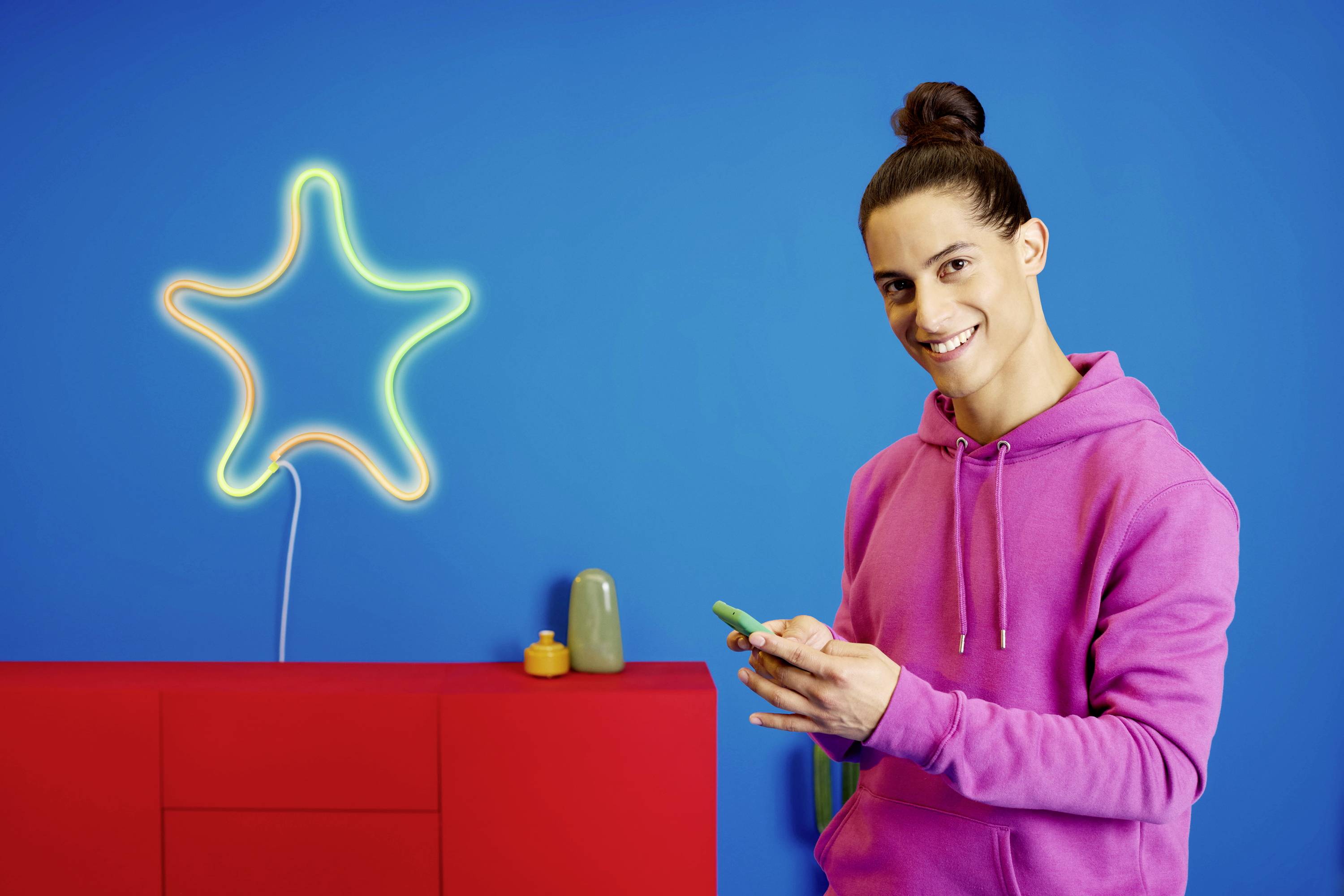 A smiling person in a pink hoodie is holding a smartphone. In the background, a glowing star decoration is displayed against a blue wall.