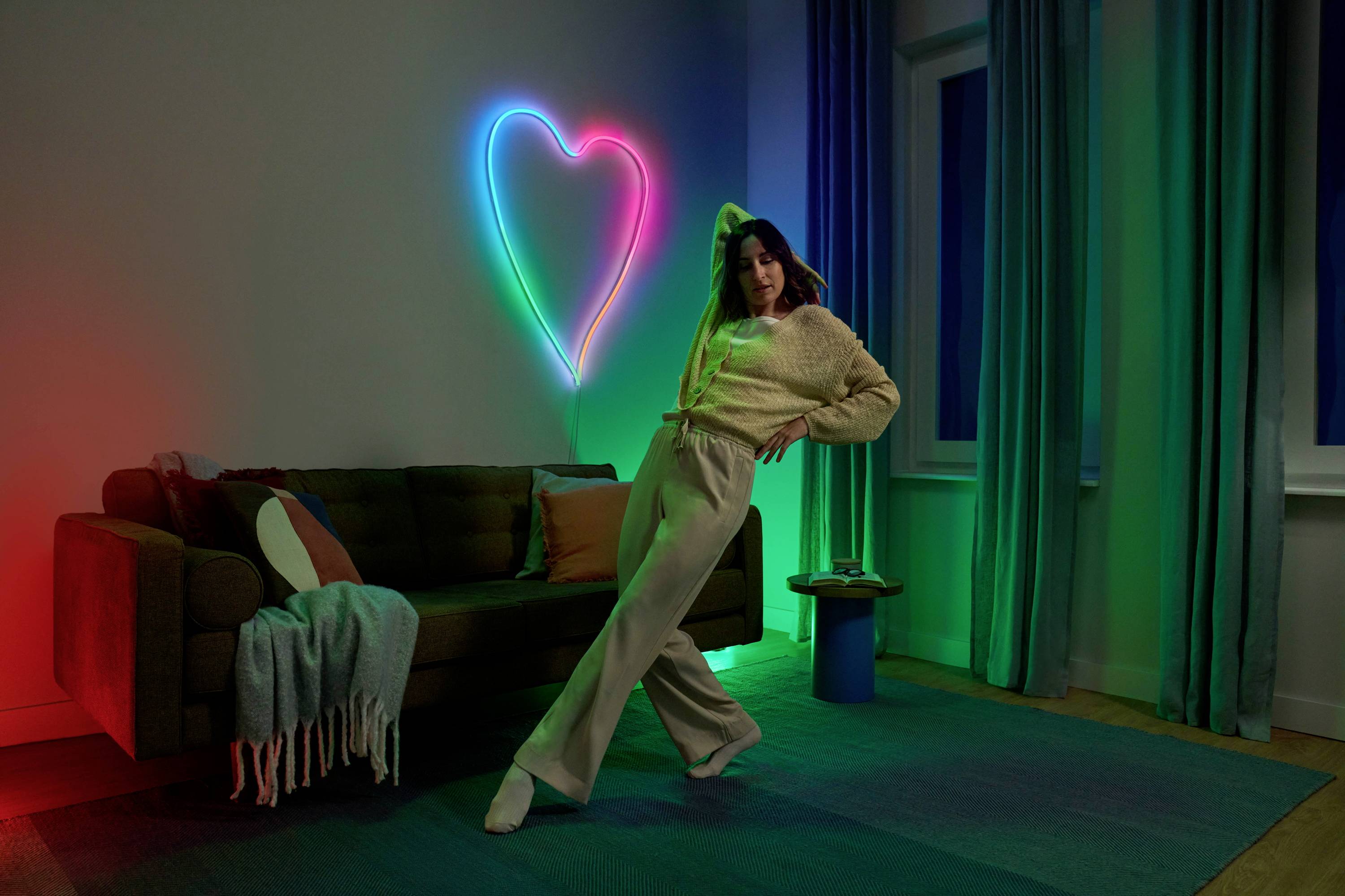 A woman poses in the living room with a neon heart on the wall, surrounded by coloured light. Sofa with a throw and cushions, curtains at the window.