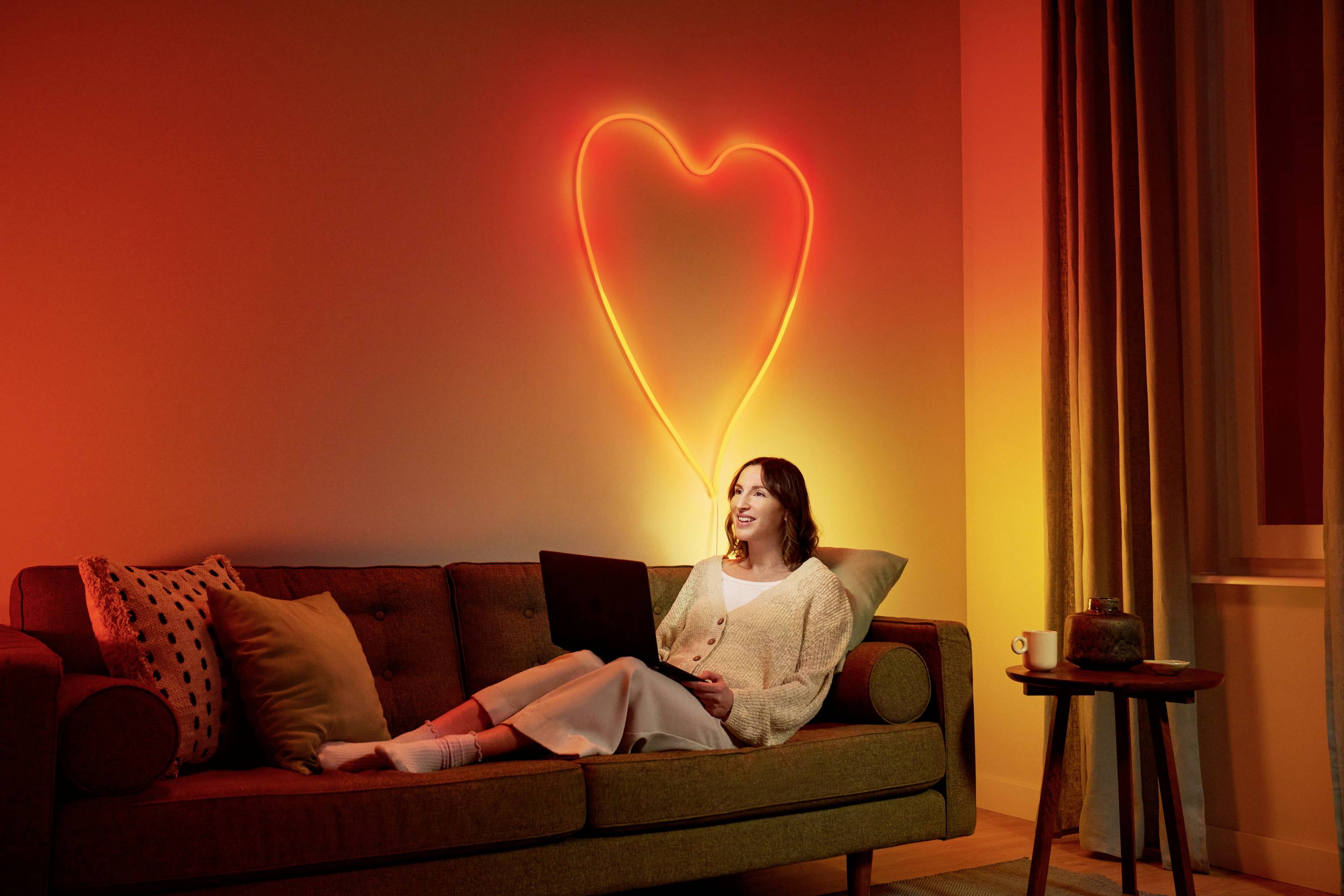 A woman sits smiling with a laptop on a sofa, surrounded by cosy lighting. A neon-coloured heart glows on the wall.