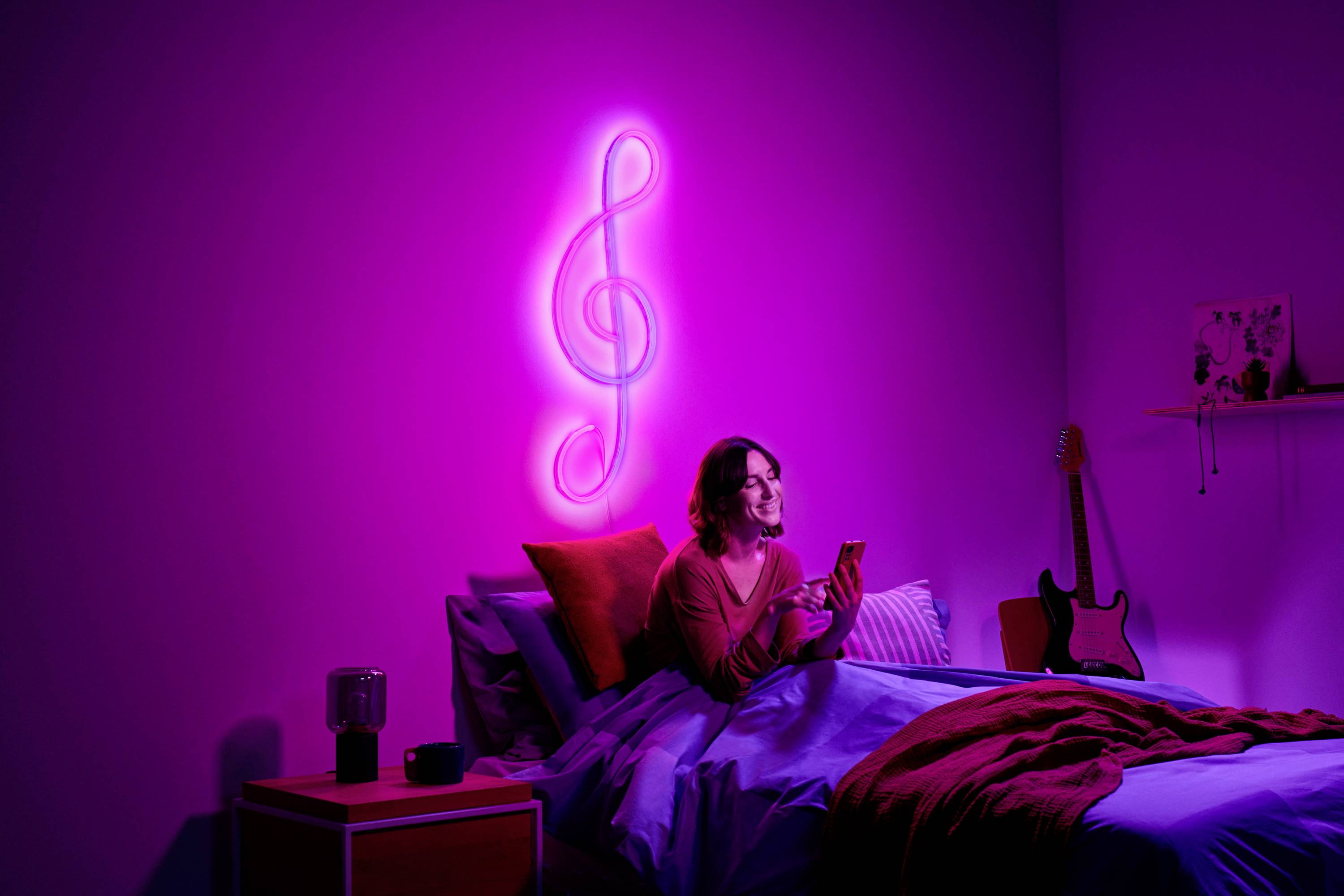 A person is sitting on a bed in a pink-lit room, holding a smartphone. A glowing treble clef hangs on the wall.