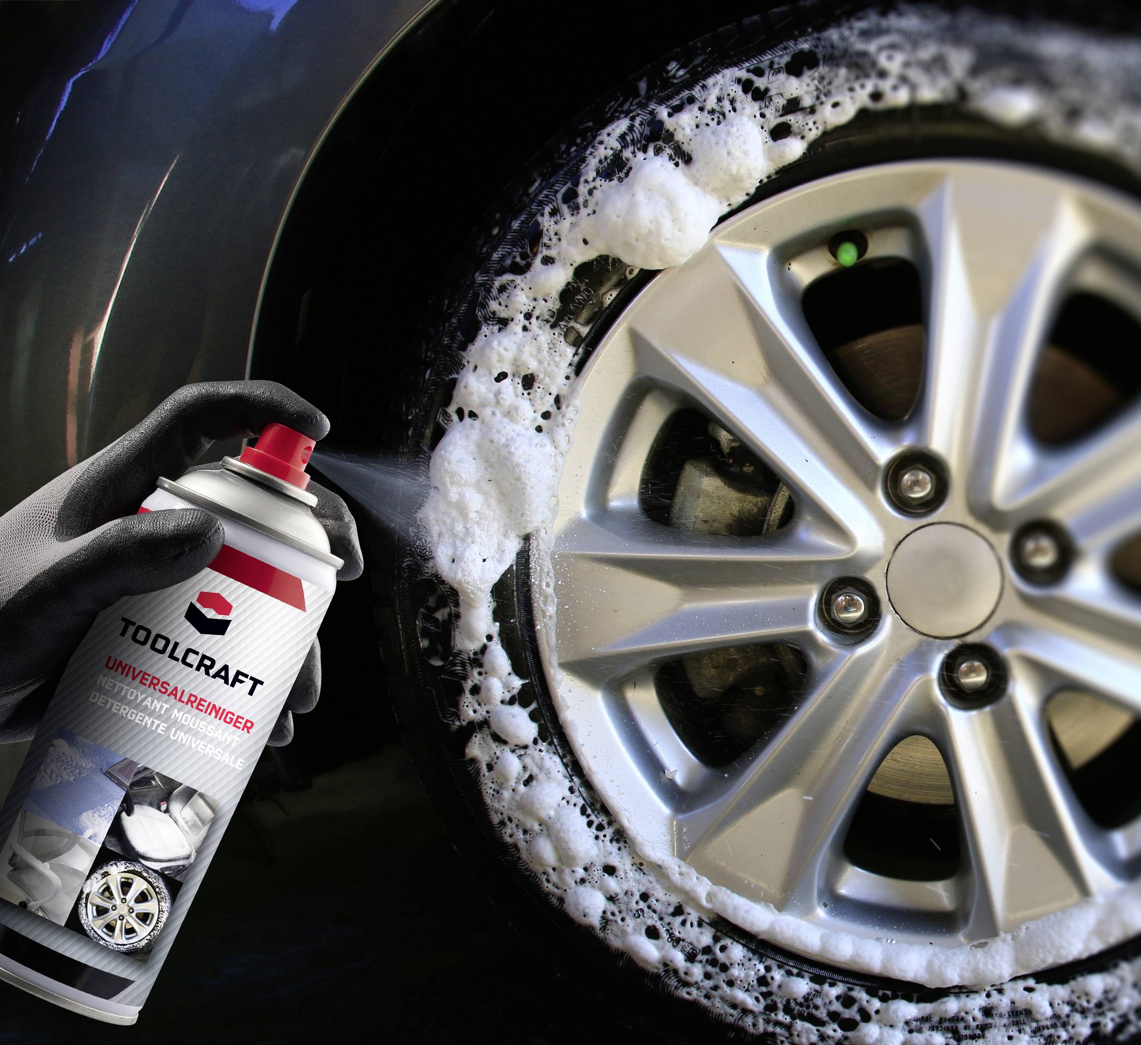 A hand sprays cleaner onto a wet car tyre, which is covered in foam. The cleaner is from the Toolcraft brand.