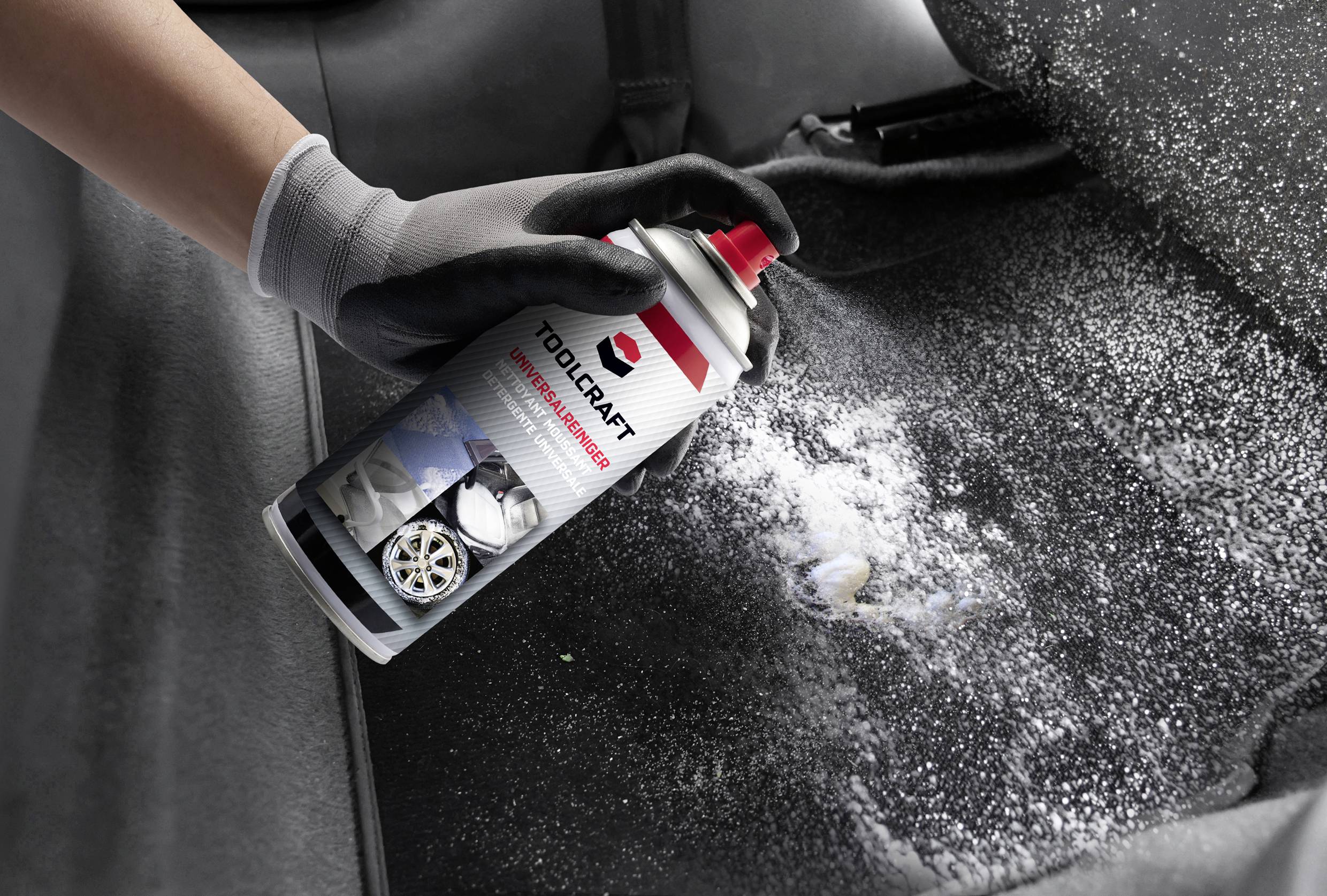 A person wearing a glove is spraying foam cleaner onto a car carpet to clean it.