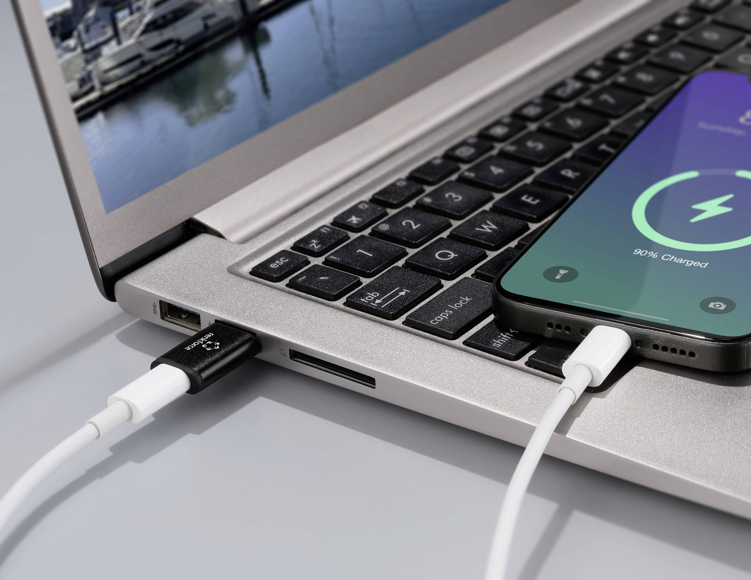 A smartphone is connected to a laptop via a cable and displays '90% charged'. Boats can be seen in the background.