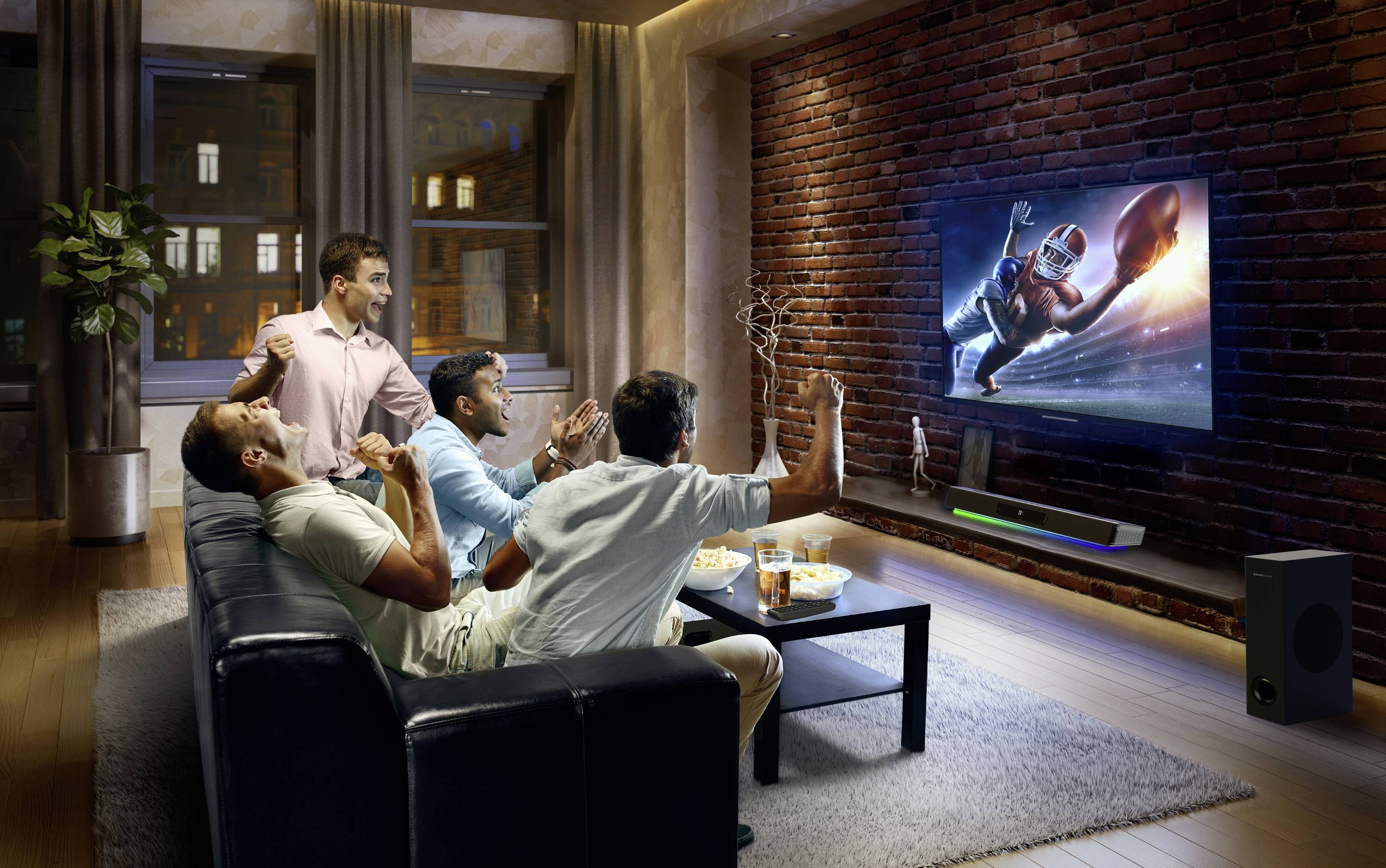 Four men are cheering on a sofa while watching an American football match on the telly. Snacks are laid out on a table.