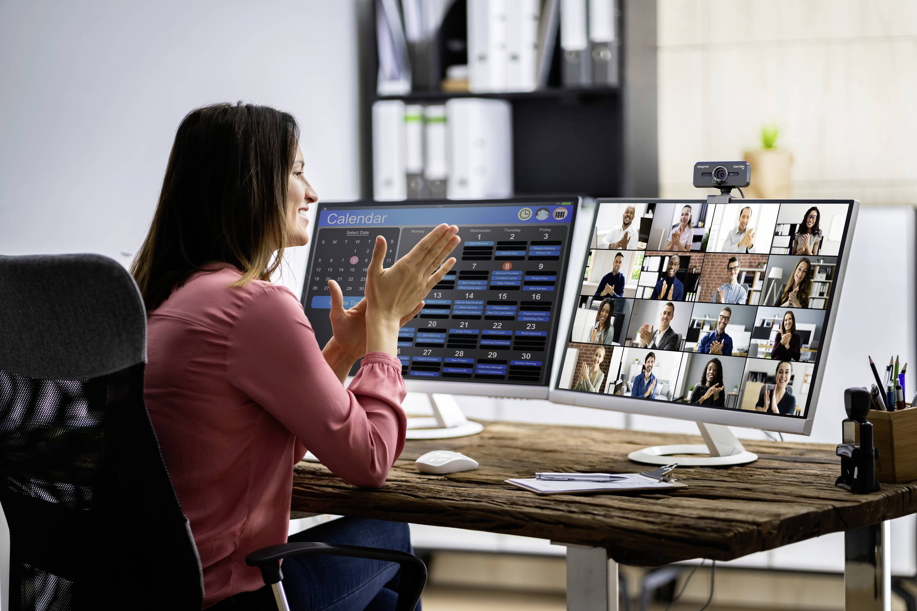 A woman is sitting at a desk and participating in a video conference with multiple participants on two monitors.