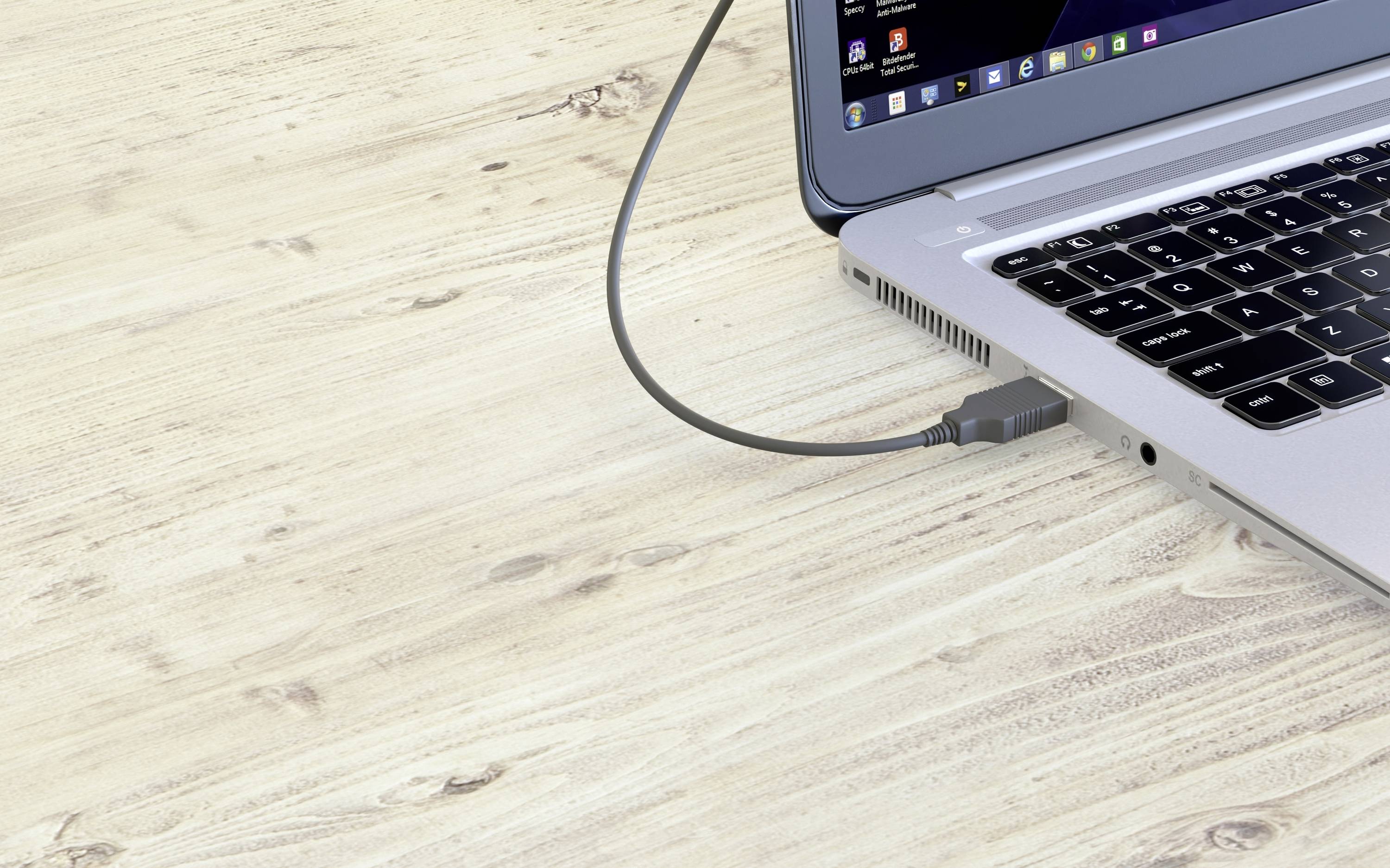 A laptop on a wooden table has a plugged-in USB cable. The screen is partially visible and shows open programmes.