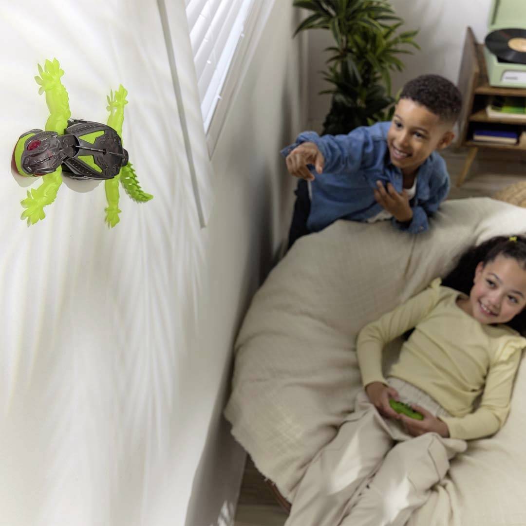 A boy eagerly guides a toy wall crawler on the wall, while a girl sits on the ground watching.