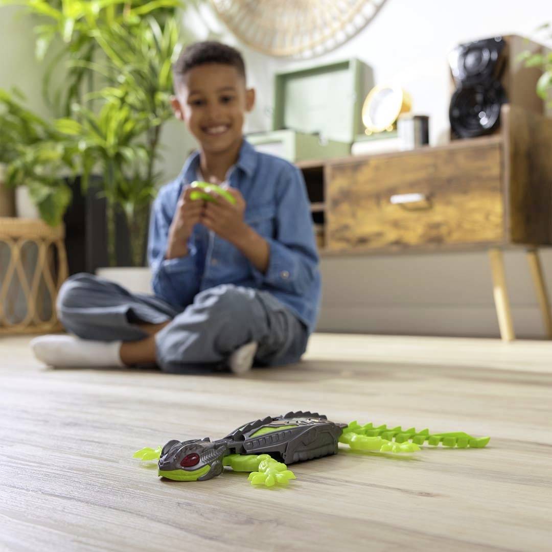 A boy sits smiling on the floor in a living room, playing with a toy dinosaur that has glowing parts.