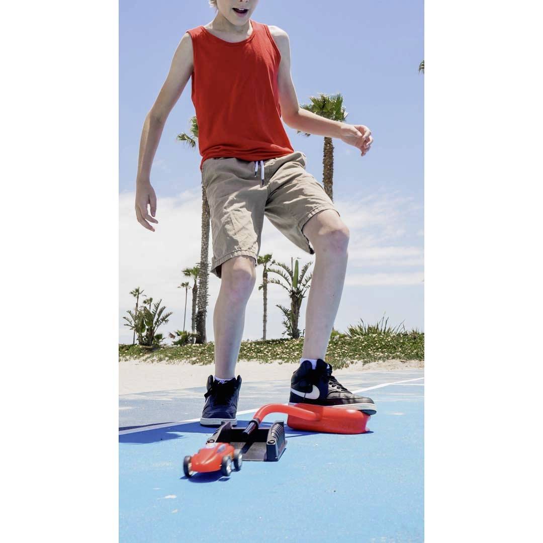 A child in a red vest and beige shorts is standing on a blue surface near palm trees. A skateboard is positioned in front of them.