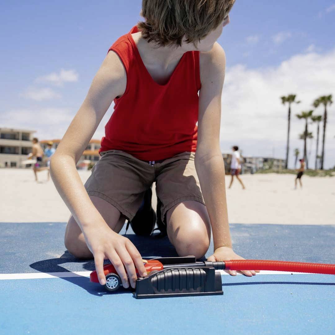 A child in a red T-shirt and shorts is playing with a red pump on a blue floor at the beach. Palm trees and buildings are visible in the background.