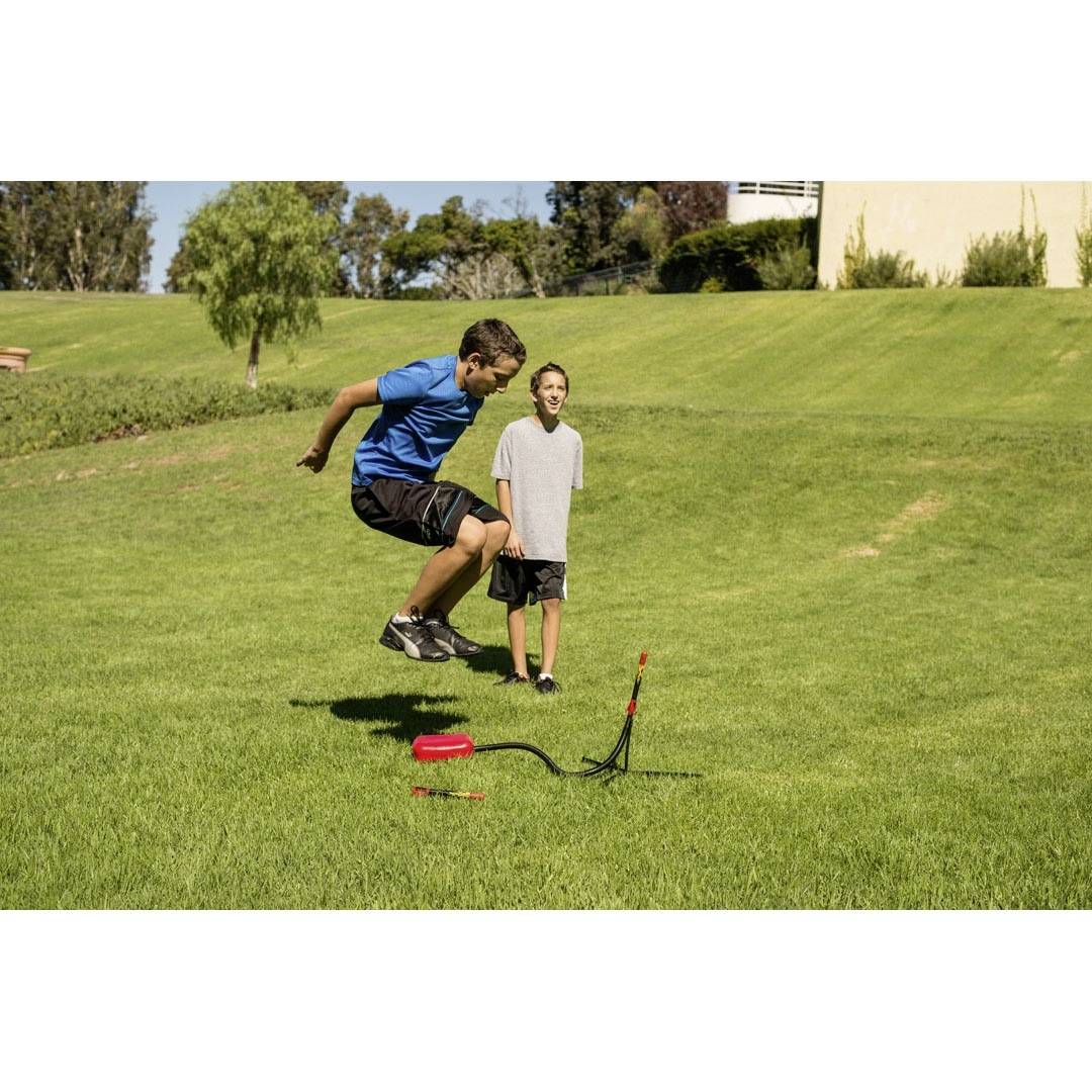 Two boys are playing on a meadow. One jumps into the air whilst the other watches. A toy rocket is set up on the ground.