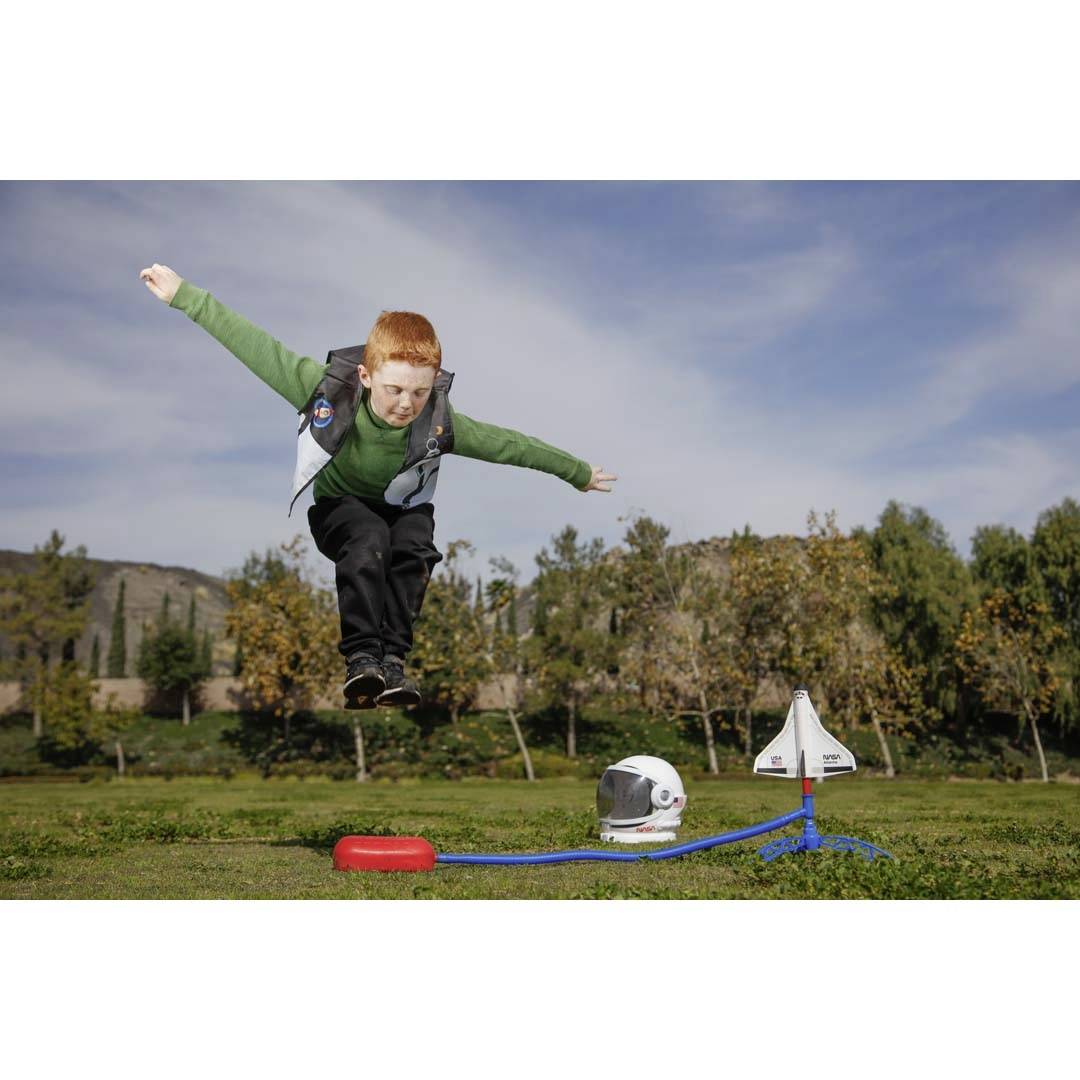 A boy jumps joyfully into the air next to a toy rocket set with a helmet outdoors. Trees and a blue sky are visible in the background.