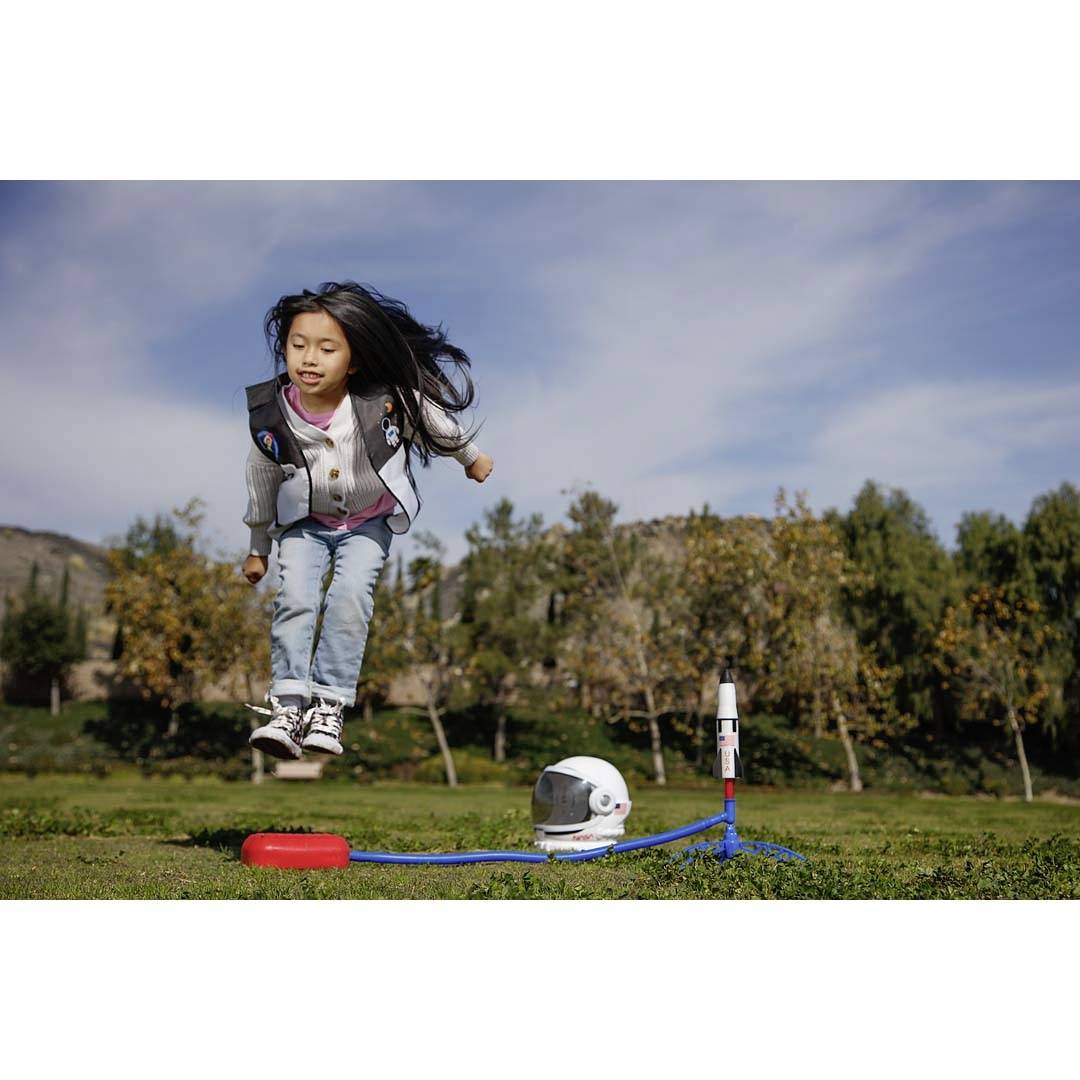 A child jumps into the air in a park, next to a toy rocket and an astronaut's helmet on the grass.