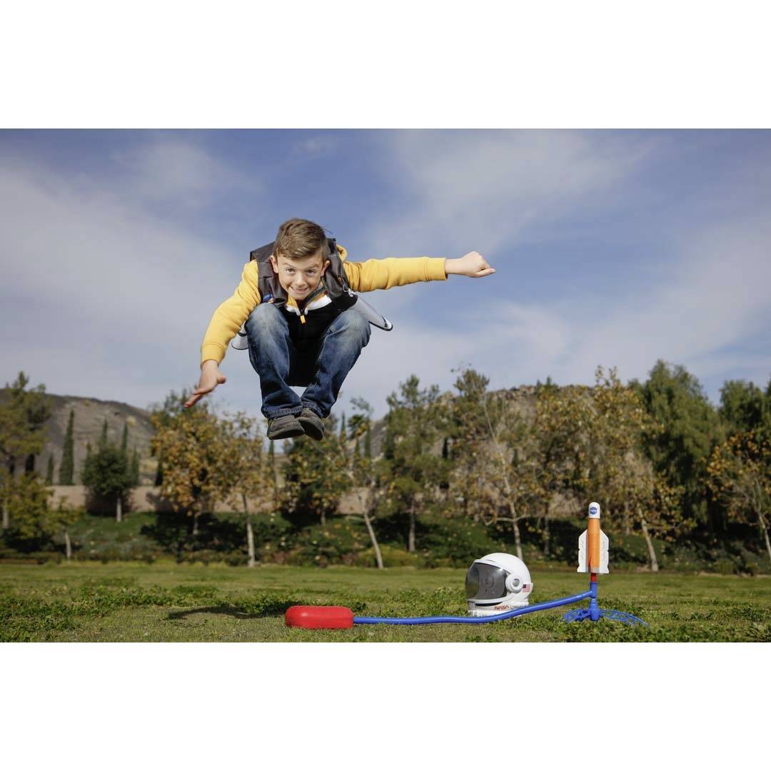 A boy jumps in a park, with a toy rocket and an astronaut's helmet lying on the ground beside him.