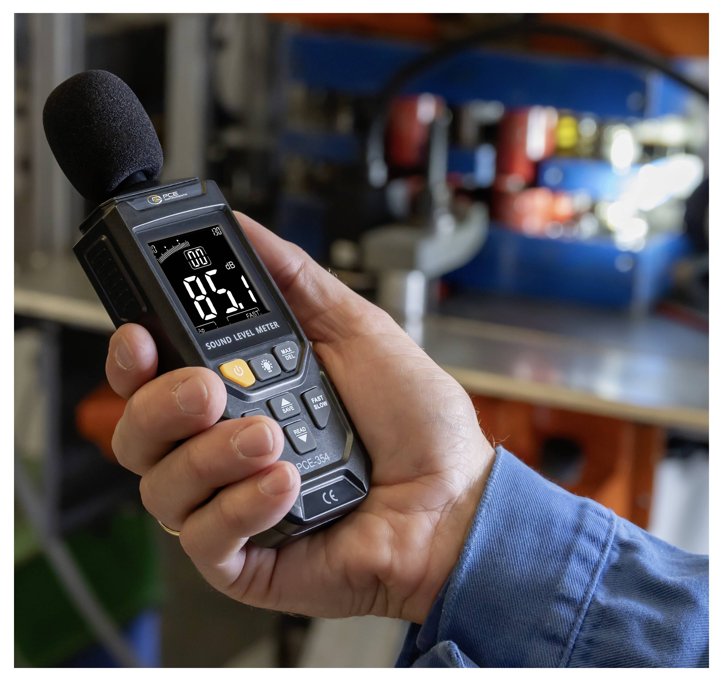 Hand holding a sound level meter with foam microphone, reading 85.1 dB, in a workshop or industrial setting.
